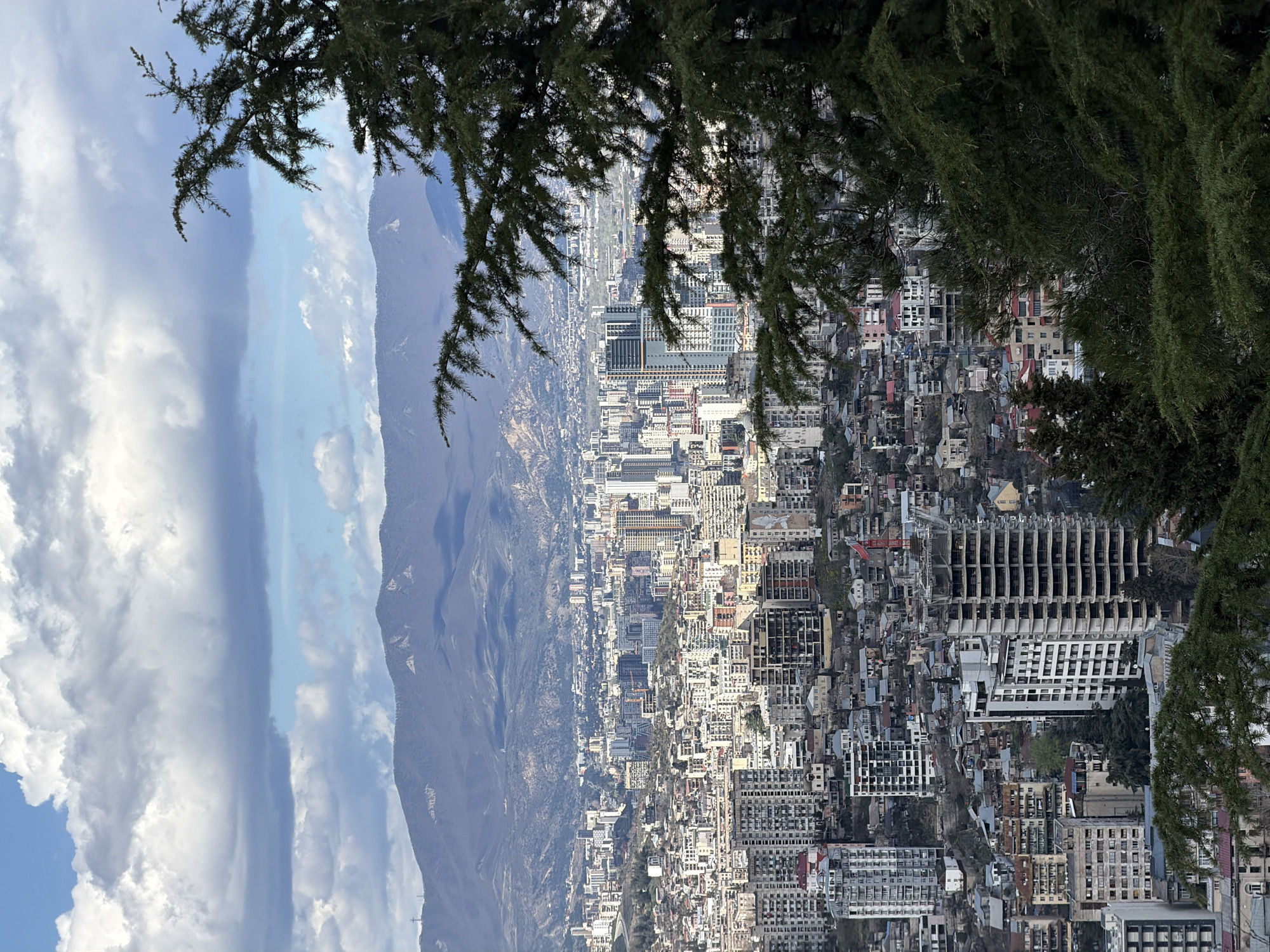 From an elevated position, a panoramic view encompasses a city, likely Tbilisi, Georgia, nestled between mountains and a sky adorned with scattered clouds. The foreground features the foliage of trees, framing the urban landscape. The city is characterized by a mix of architectural styles, with buildings of varying heights and designs densely packed together. Mountains rise in the background, providing a scenic backdrop. The lighting suggests a daytime scene, with sunlight illuminating the city and casting shadows on the mountains.