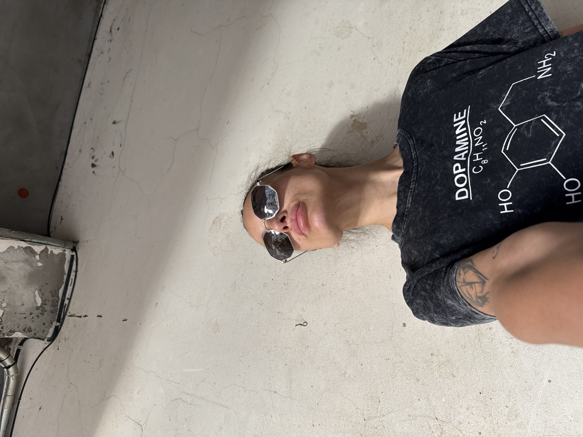 A person lies on a light-colored, possibly concrete, surface with visible cracks and imperfections. The person is wearing sunglasses and a dark T-shirt with the word "DOPAMINE" and a chemical formula printed on it. The T-shirt appears to be acid-washed or have a similar worn effect. A partial view of an arm is visible, bearing a tattoo. The location is unidentified, and there is no evidence to suggest that the image was taken in Pleven, Bulgaria. A corner of a gray wall or structure is visible on the left side of the frame, showing piping and a possible drainage feature.