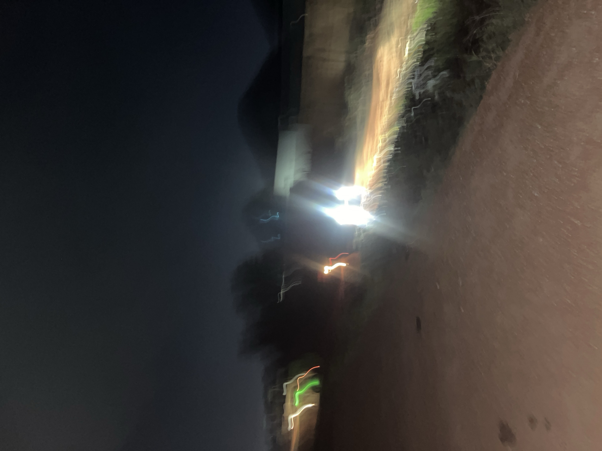The image shows a blurry, low-light scene, possibly taken at night, with a focus on a light source. To the right, what appears to be a dirt road or path is visible. Some vegetation is seen along the side of the path. The background is dark and indistinct. The location is unknown, and Abuja, Nigeria, cannot be confirmed. No people, actions, or interactions are clearly discernible.