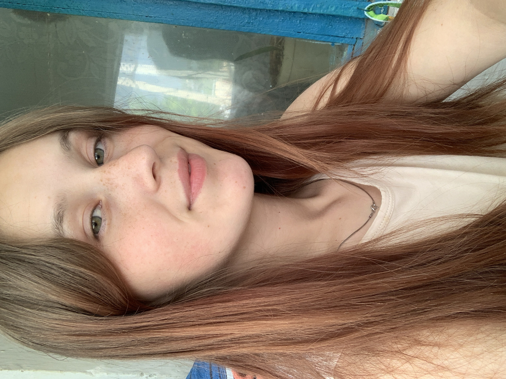 A close-up, brightly lit portrait features a young woman with long, reddish-brown hair parted on the side. She has fair skin, adorned with numerous freckles across her nose and cheeks. Her light-colored eyes gaze directly at the viewer, and a gentle, subtle smile plays on her light pink lips. She is wearing a simple, light-colored (possibly cream or white) sleeveless top, and a delicate silver chain necklace with a small pendant is visible around her neck.
Her hair flows extensively, framing her face and covering parts of the lower and right sides of the image. The background is softly blurred, revealing reflections of outdoor elements, such as foliage or sky, in what appears to be a window or glass surface. A prominent, textured blue wooden structure stretches across the upper part of the background, possibly indicating a window frame or architectural detail. The natural light suggests a daytime setting. The overall expression conveyed is calm and pleasant. No text is visible in the image.