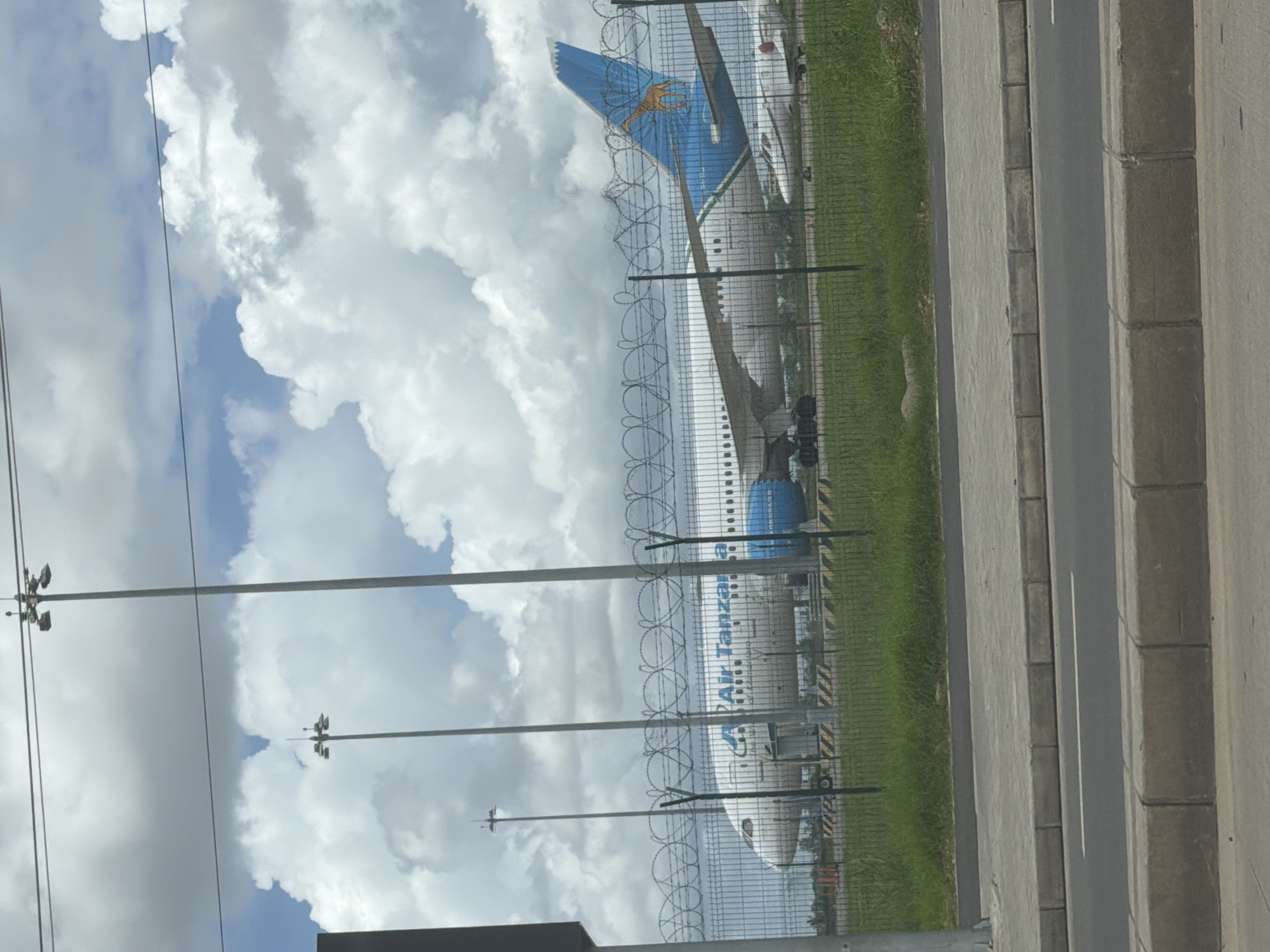 An Air Tanzania passenger jet is behind a security fence. The city is unknown. The sky is cloudy. There is grass and concrete near the fence. There are light poles and wires.