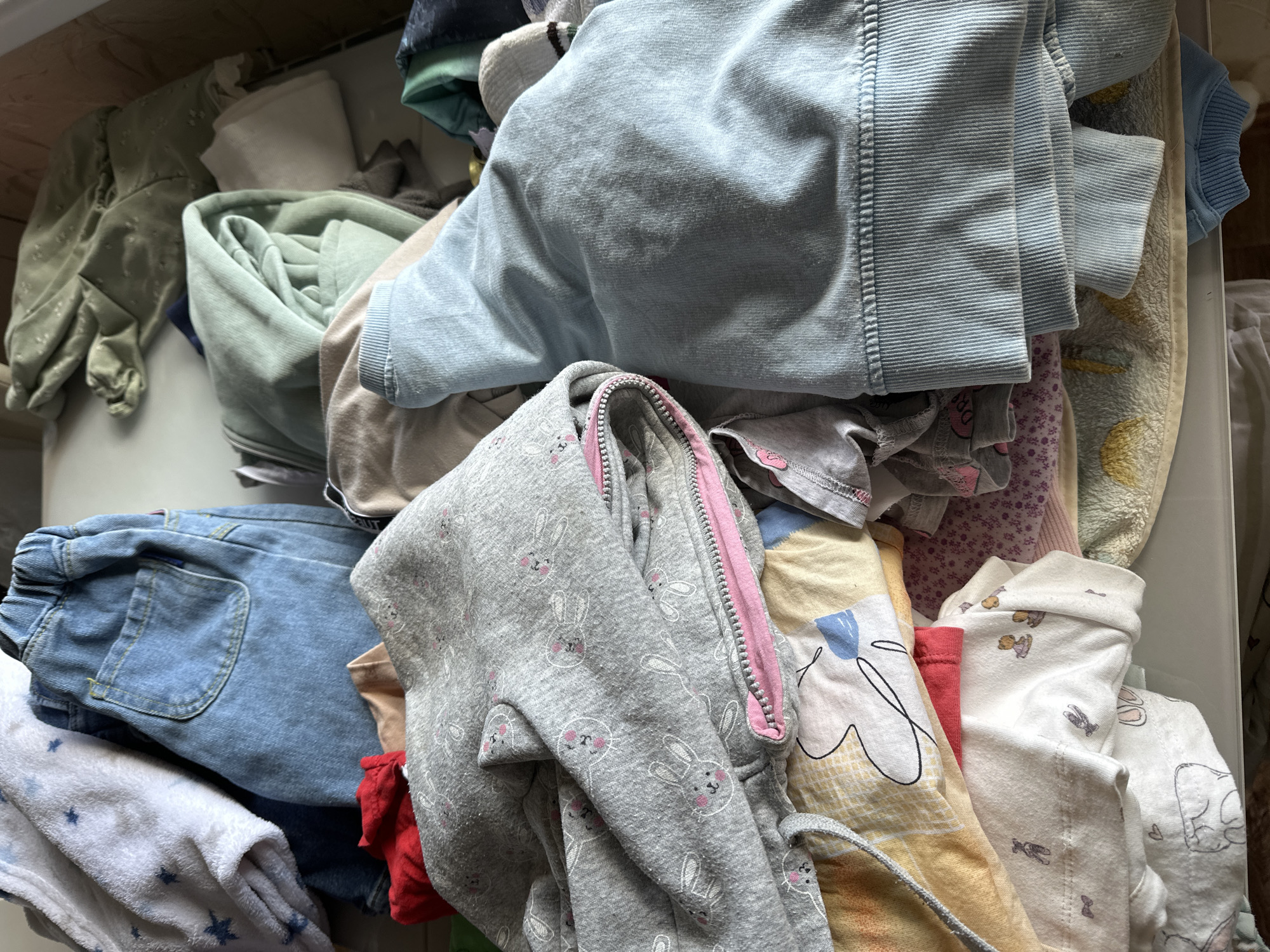 A high-angle close-up photograph shows a pile of folded and crumpled children's clothing in an indoor setting. The clothing includes a light blue corduroy jacket, a gray zip-up hoodie patterned with bunny faces and pink trim, blue jeans, and several other articles of clothing with various patterns and colors. The location cannot be determined.