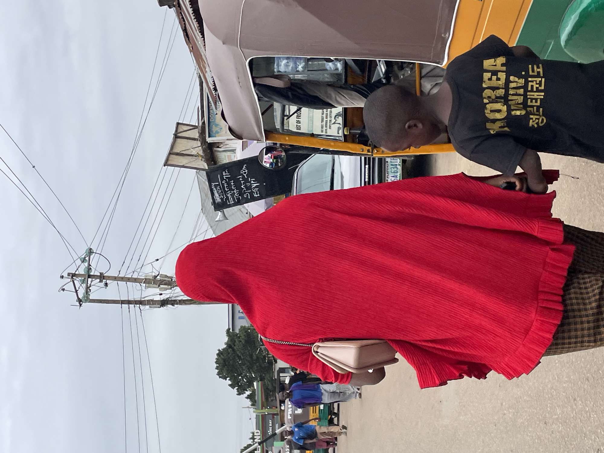 The image captures a street scene in Jos, Nigeria. A woman wearing a red, hooded garment and a small purse is partially visible. A young boy in a black shirt with the text "KOREA UNIV." and Korean script stands next to her, appearing to interact with something off-camera. A yellow auto rickshaw, commonly used for transportation in the area, is parked nearby. A sign attached to the rickshaw displays handwritten text including countries like "ENGLAND," "SPAIN," and "EGYPT," and the date "31/3/21." Above, a tangle of electrical wires stretches across the sky. Other individuals are visible in the background. A sign reads "BE BAY" and "DOWNTO LAND." A car license plate is partially visible, reading "SA-634TF."