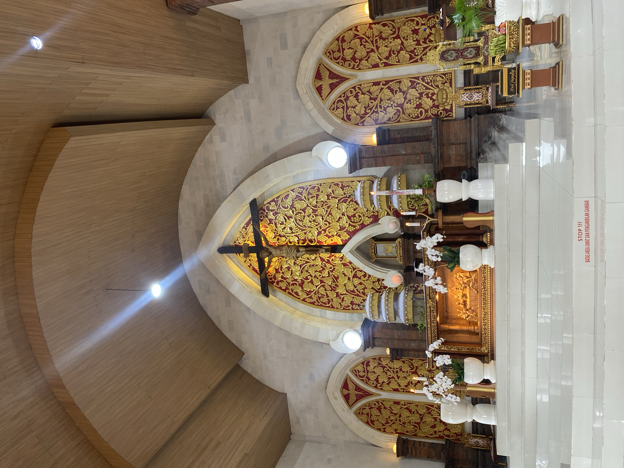 The image captures the interior of a church, likely located in North Kuta, Indonesia, judging by the architectural style and religious symbols. The focal point is a crucifix mounted against an ornate, gold-and-red patterned background. The ceiling is partially visible, constructed from light wood panels, and includes a light fixture casting a beam of light. Below the crucifix is the altar area, featuring decorative elements, candles, and white vases with flowers. The altar is elevated by white steps. A sign written in Indonesian, including the word "STOP," is visible on the steps.
