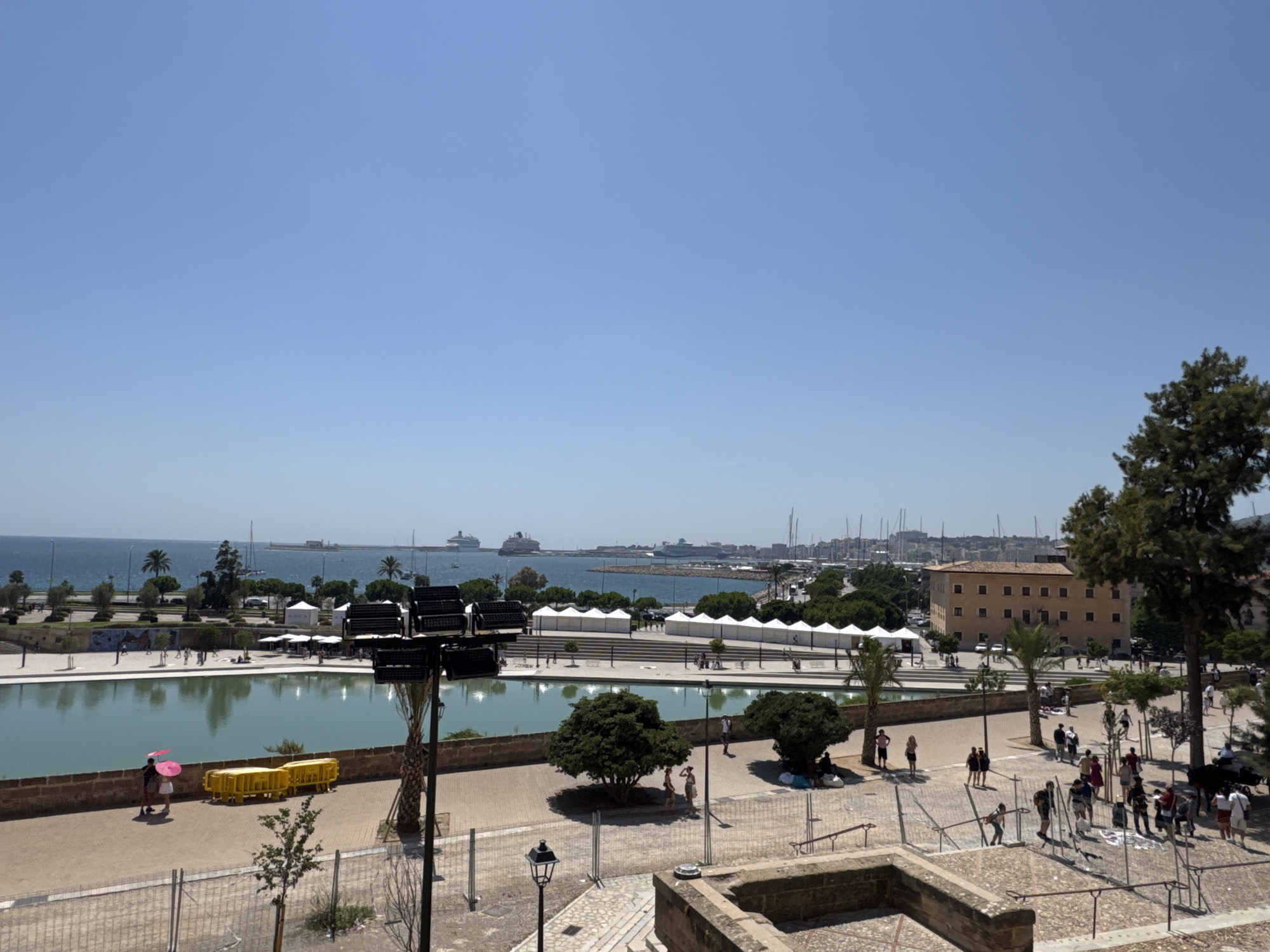The image shows a scenic view of Palma de Mallorca, Spain, on a clear, sunny day. In the foreground, there is a large, rectangular pool of calm, blue water. A stone pathway runs along the edge of the pool, with a few people walking on it. To the left of the pool, a couple is walking together, one holding a pink umbrella. There are also some yellow barriers placed along the path.
Beyond the pool, there is a promenade with several white tents set up, suggesting an outdoor market or event. Palm trees and other greenery are scattered throughout the area. In the background, the city's harbor is visible, filled with numerous sailboats and a few large cruise ships. The city skyline features buildings of various sizes and architectural styles, including a prominent tan building with many windows. The deep blue sea stretches to the horizon under a vast, clear blue sky. The overall scene depicts a vibrant and picturesque waterfront area in Palma.