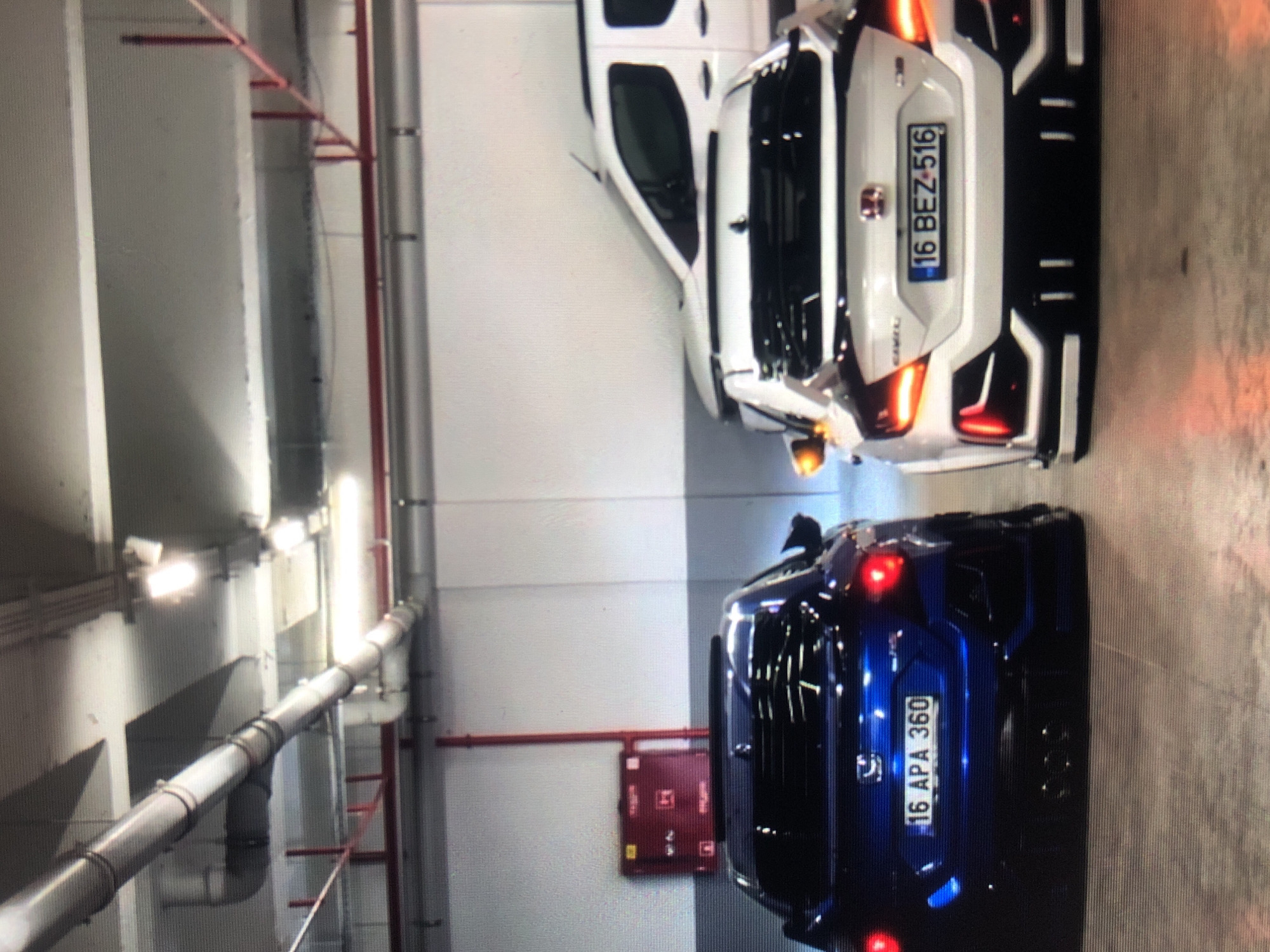 The image shows two cars parked in an indoor parking garage. A white van is parked next to a white car with license plate "16 BEZ 516." A dark blue car is parked facing the opposite direction with license plate "16 APA 360". The parking garage has fluorescent lights and a red fire extinguisher box on the wall. There are pipes and support structures running along the ceiling. The location cannot be determined from the image.