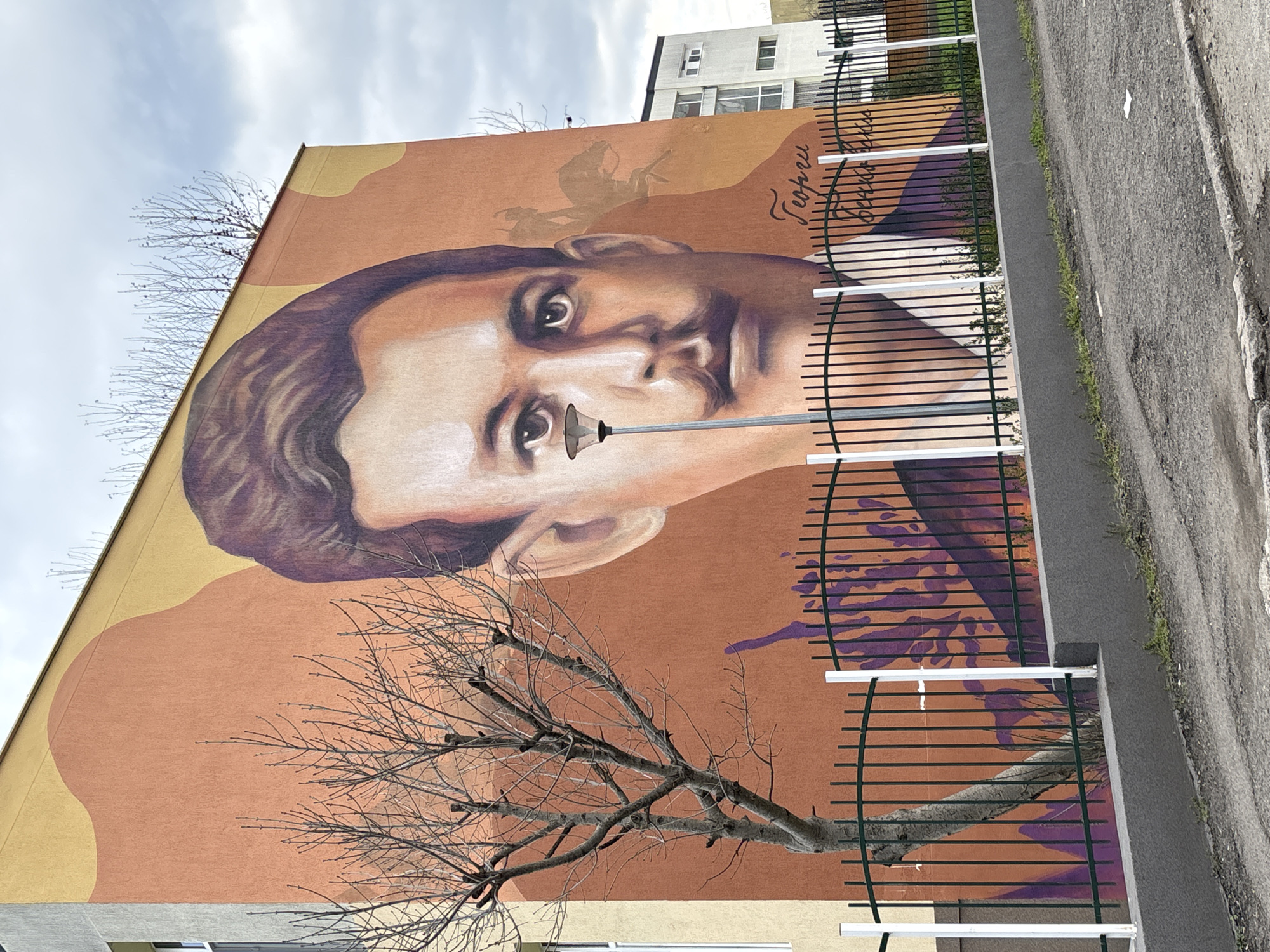 A mural of a man's face, with the name "Георги Бенковски" in Cyrillic script painted near the upper right, covers the side of a building in Burgas, Bulgaria. A leafless tree is partially in front of the mural. A dark green, curved-top metal fence with white vertical posts runs along the base of the building, adjacent to a gray asphalt sidewalk and street. The sky is overcast. Another building is visible behind the top of the mural. A streetlight or street sign extends from the man's nose in the mural.