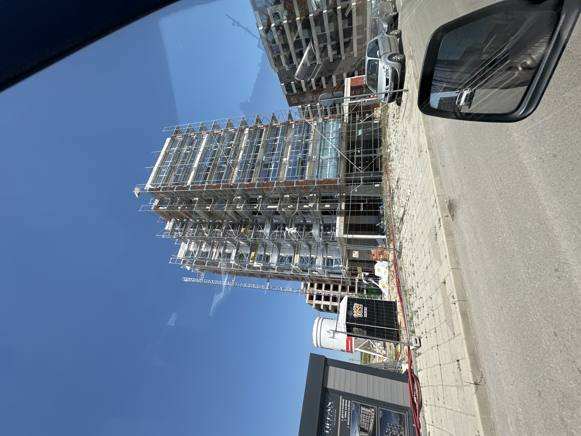 The image shows a construction site in a city with a tall, multi-story building covered in scaffolding. The building has exposed brick and glass windows. In the foreground, there is a sidewalk with a curb, and a street. There are two cars parked on the side of the street. To the left of the building, there is a small temporary structure with signage that reads "TIFFAN". There is also a portable toilet and a large white silo. The sky is clear and blue. The image appears to have been taken from inside a vehicle, as evidenced by the reflection in the side-view mirror.