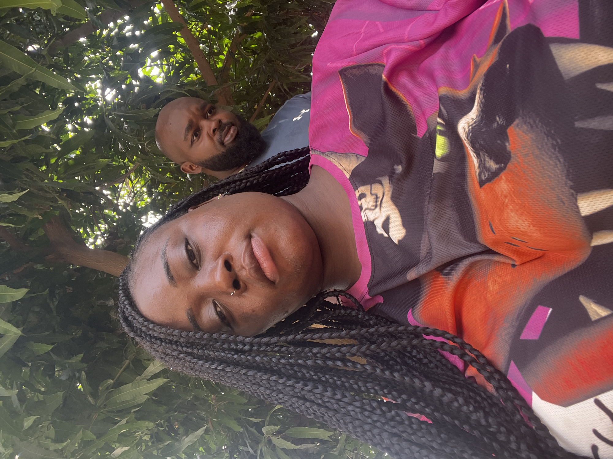 The image captures two individuals, a woman and a man, from a low-angle perspective looking up into a dense canopy of green leaves. The overall scene suggests an outdoor, natural setting during daylight hours, likely in Kubwa, Nigeria.
In the foreground, a woman with dark skin and long, black braided hair is prominent. She has a small silver nose piercing on her left nostril and full lips. She is wearing a long-sleeved top with a vibrant, abstract pattern in shades of hot pink, black, orange, and purple. Her expression is calm and she is looking directly towards the camera.
Behind and slightly to the left of the woman is a man with dark skin, a bald head, and a well-groomed dark beard. He is wearing a plain dark grey or blue t-shirt. He also looks directly towards the camera with a neutral expression.
The background consists entirely of lush green foliage from a tree, with bright sunlight dappling through the leaves, indicating a sunny day. No text is visible in the image. The composition suggests a casual selfie or photograph taken from below, possibly with the subjects lying down or looking up.