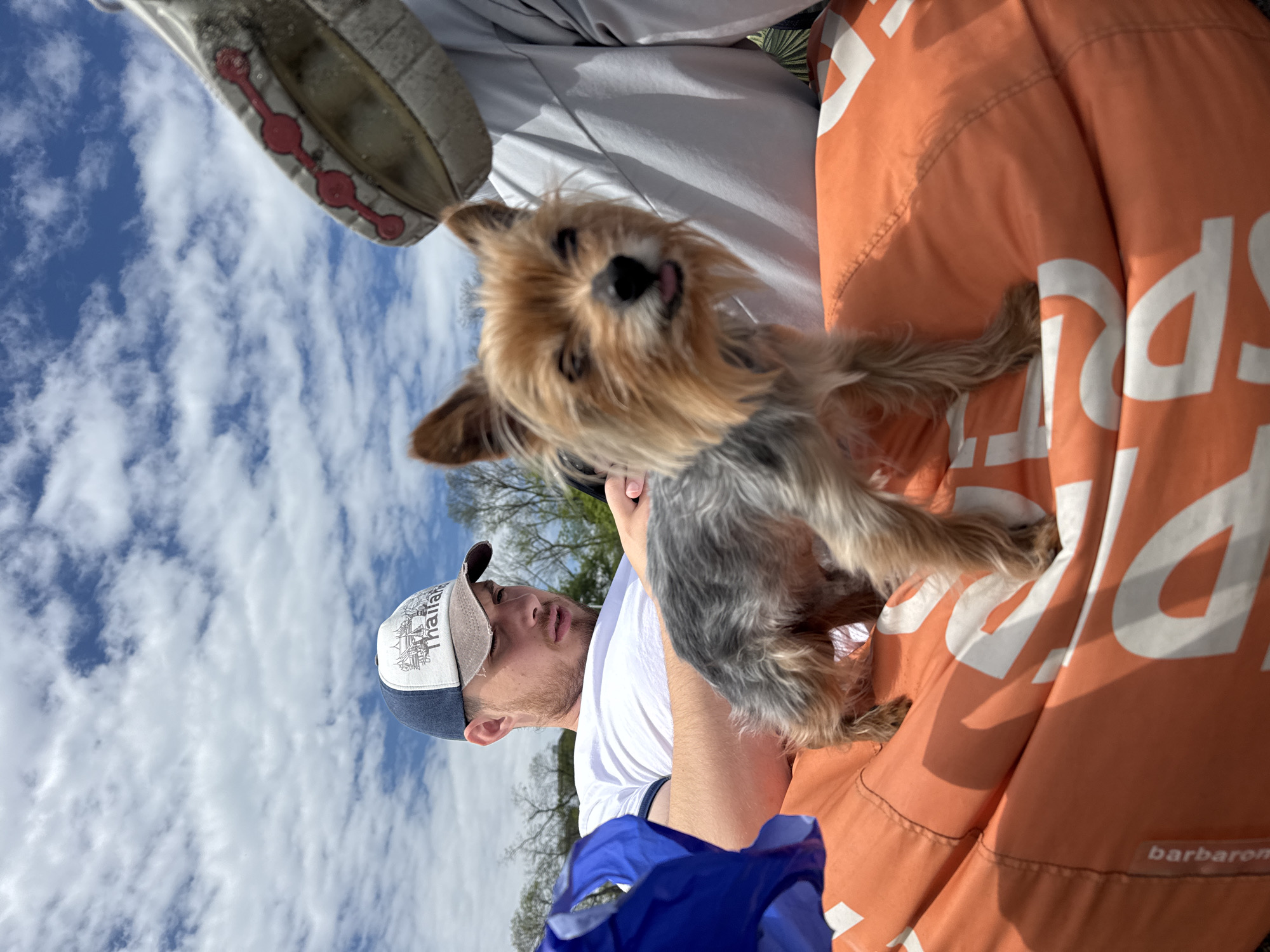 A man is seated outdoors, cradling a small dog in his lap. He is wearing a white t-shirt and a baseball cap with the word "Thailand" embroidered on it. The dog is a Yorkshire Terrier with tan and grey fur. The man is holding the dog with his left hand, and the dog's tongue is slightly visible. The man and dog are sitting on an orange bean bag chair with the brand name "barbaron" printed on it. To the left of the man, a shoe sole is visible, implying another person is nearby. The sky is partly cloudy with blue patches. It is not possible to identify the location as Burgas, Bulgaria, without additional information.