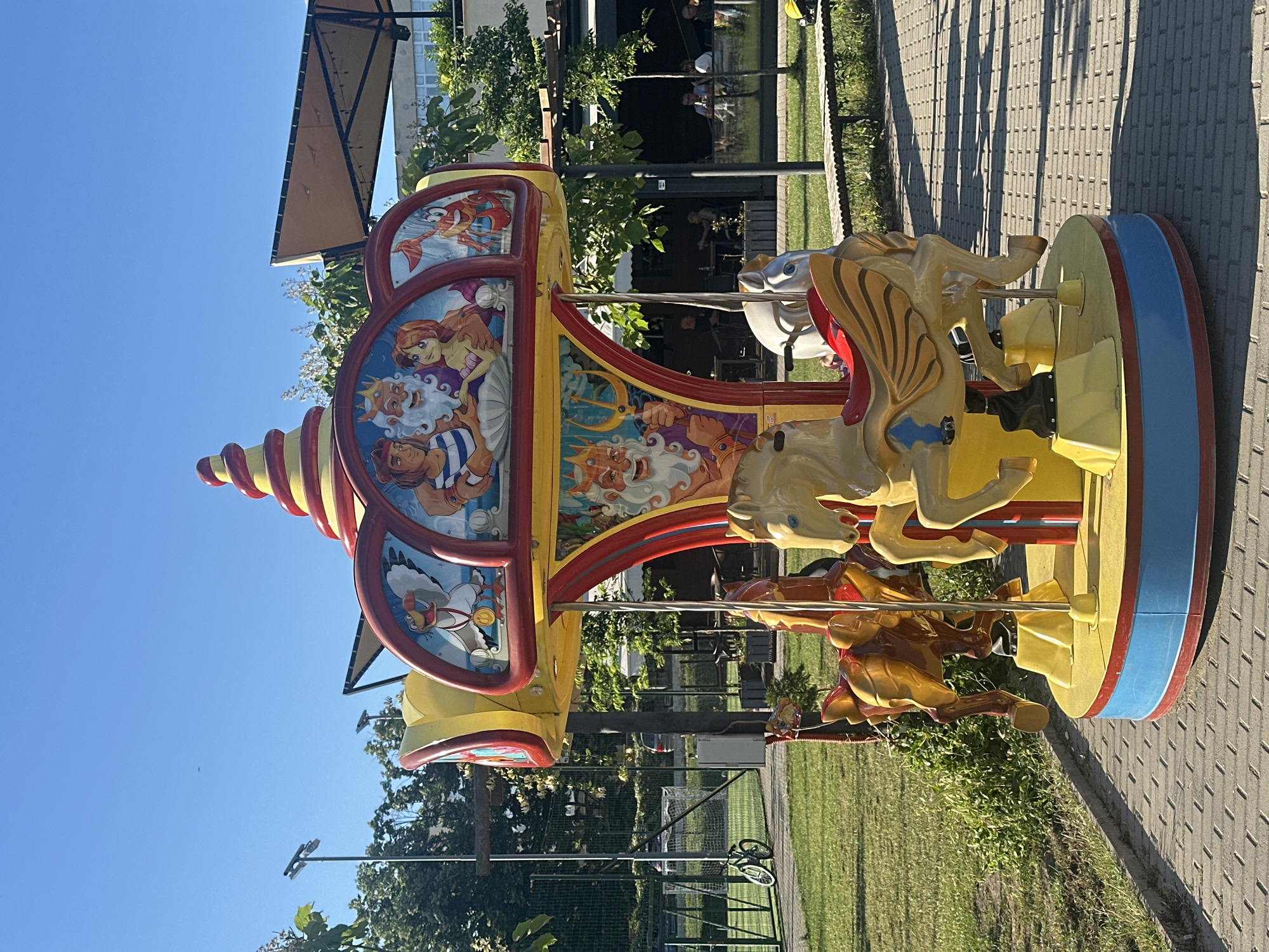 The image shows a merry-go-round in an outdoor setting. The merry-go-round is brightly colored, with yellow horses and a red and yellow striped top. The sides of the merry-go-round are decorated with cartoon images, possibly from a fairy tale or children's story, featuring figures with crowns, beards, and mermaids. There are also images of a crab and a white bird. The merry-go-round is on a paved area with grass and trees surrounding it. In the background, there are buildings and a clear blue sky, suggesting it is daytime and the weather is fair. The scene appears to be a park or amusement area. There is no visible text in the image.