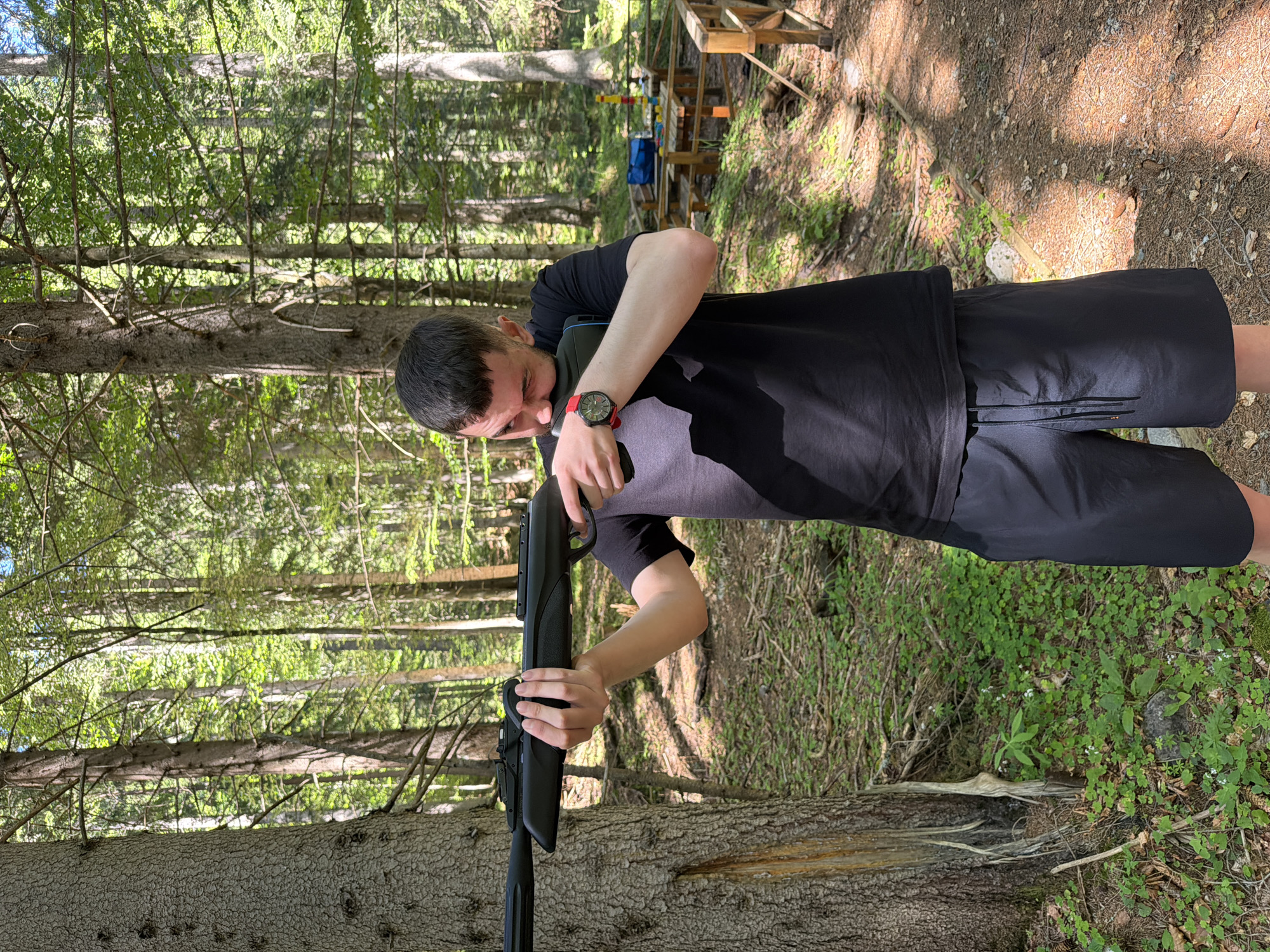 A man in a black t-shirt and black shorts is holding a black rifle in a forest. He is looking through the sights of the rifle. The forest has tall trees and green foliage. In the background, there is a wooden picnic table and a wooden fence. The ground is covered in dirt and fallen leaves. The sun is shining through the trees, creating dappled light and shadow. The scene appears to be in the city of Govedartsi, Bulgaria.