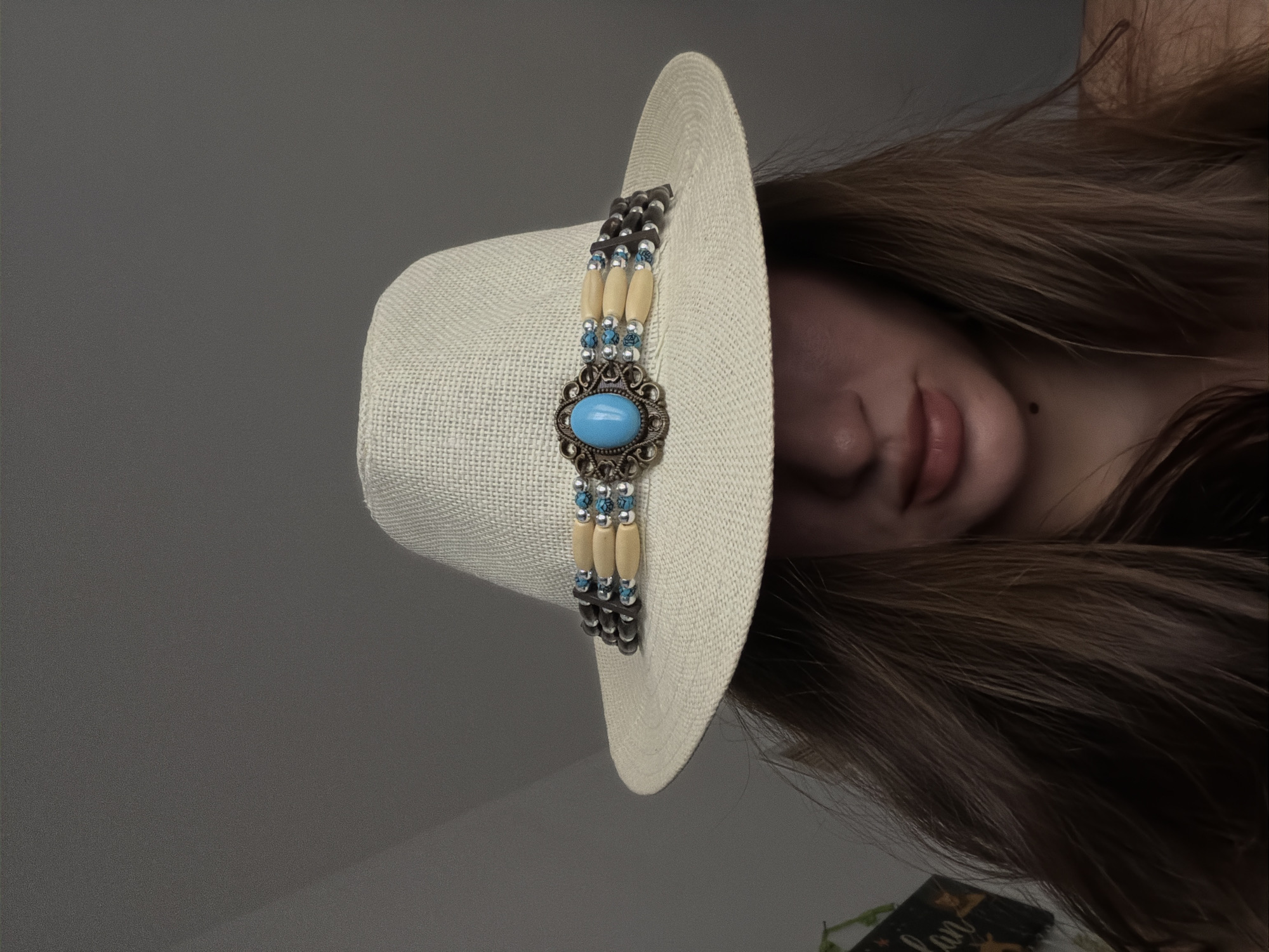 This is a headshot of a person wearing a light-colored straw hat. The hat has a decorative band featuring a central turquoise-colored oval gem surrounded by ornate metalwork and beaded elements. The person's face is partially obscured by the brim of the hat, with only their lips, chin, and lower cheeks visible. Their hair is long and brown, cascading down around their shoulders. The background is a plain, muted gray wall. There is no indication of the city of Sharm El Sheikh, Egypt, in this image.