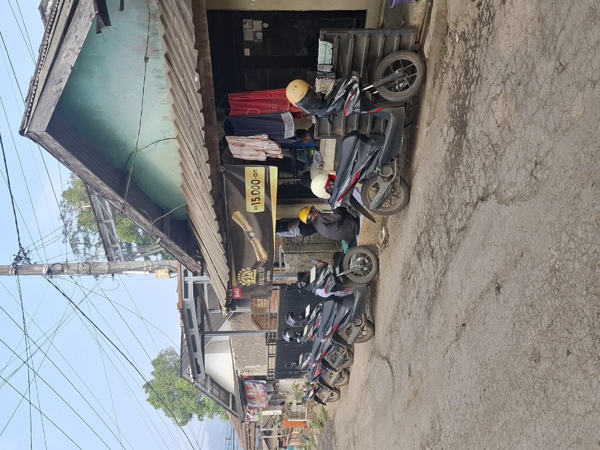 The image depicts a street scene in Ciheulang Tonggoh, Indonesia, with a row of parked motorcycles and a small building. Several black scooters are lined up on the cracked asphalt road in front of a weathered building with a corrugated metal roof. Some of the motorcycles have helmets on their seats. A banner advertising "MAGNUM KRETEK" with a price of "RP 15.000,-an" is displayed on the facade of the building. Two men are seated near the building, one wearing a yellow hard hat and the other partially visible. Clothing items, possibly for sale or display, hang on racks outside the building. The environment appears to be a residential or commercial street with overhead electrical wires crisscrossing the sky. The overall impression is one of a functional, perhaps somewhat rustic, commercial establishment in a local Indonesian community.