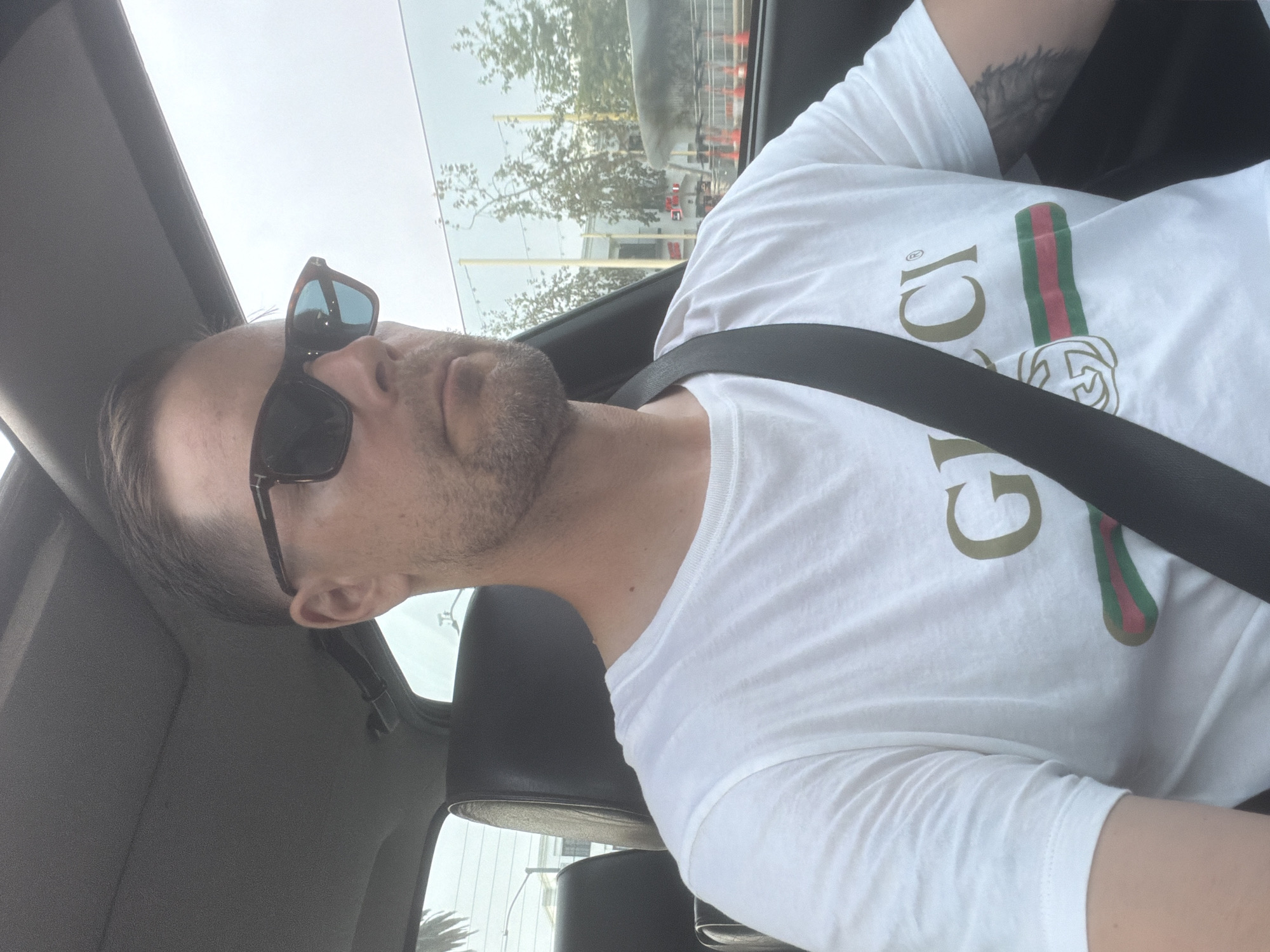 A man with short brown hair and a beard is in the driver's seat of a car. He is wearing sunglasses and a white t-shirt with the Gucci logo. The car appears to be moving, and the background shows trees and buildings in an urban environment. The photo was taken on July 28, 2023, in Santa Monica, California.
