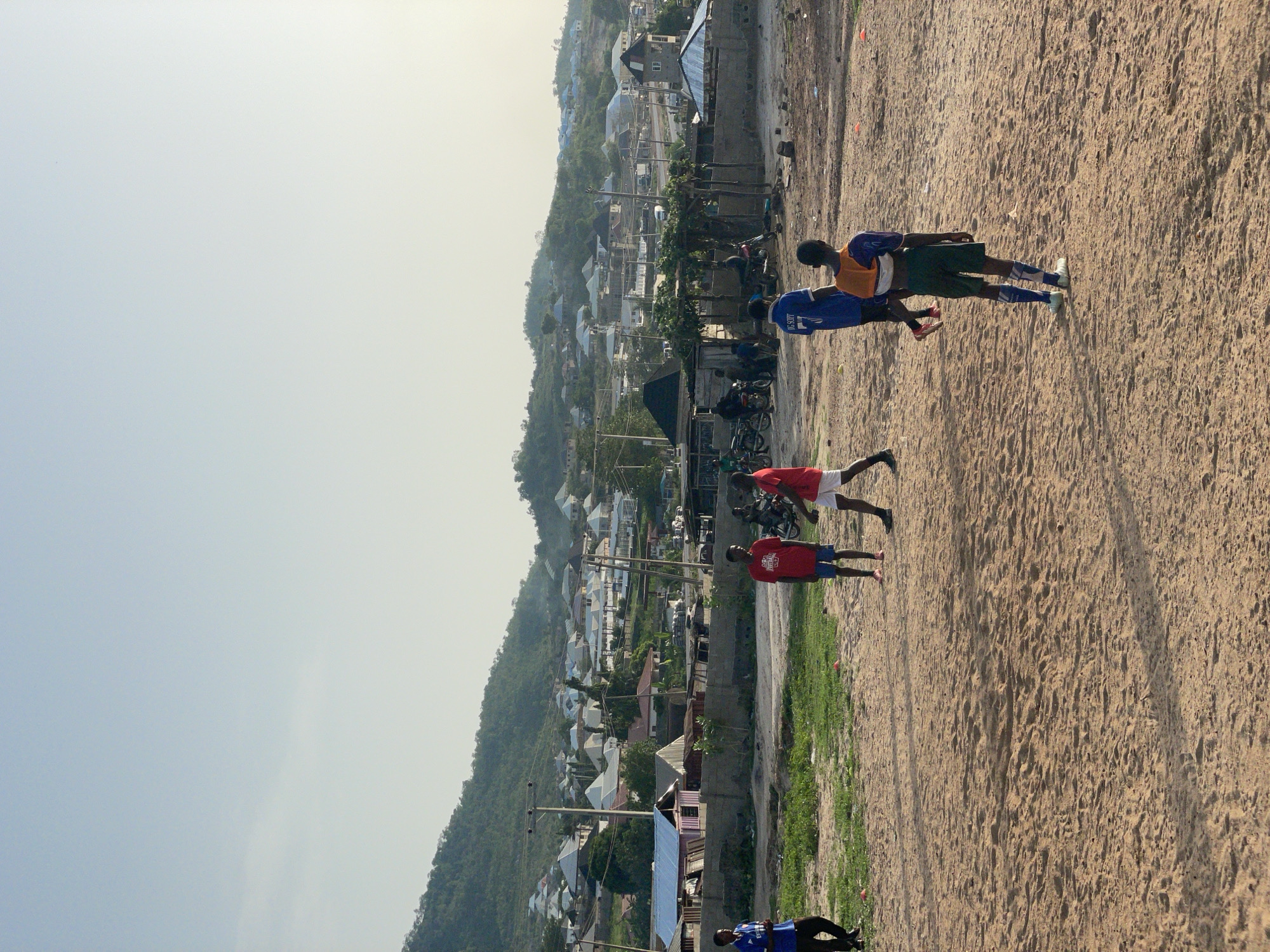 The image shows a sandy area in Lokoja, Nigeria, possibly a field, with several people. Two individuals are wearing blue jerseys, two are in red jerseys, and one is in a green jersey with an orange vest. They appear to be engaged in some form of activity, likely related to sports or training. In the background, there is a view of the city, with buildings scattered across the landscape, and a hillside or mountain. Motorcycles are parked near a wall separating the field from the city. The sky is bright, suggesting it is daytime.