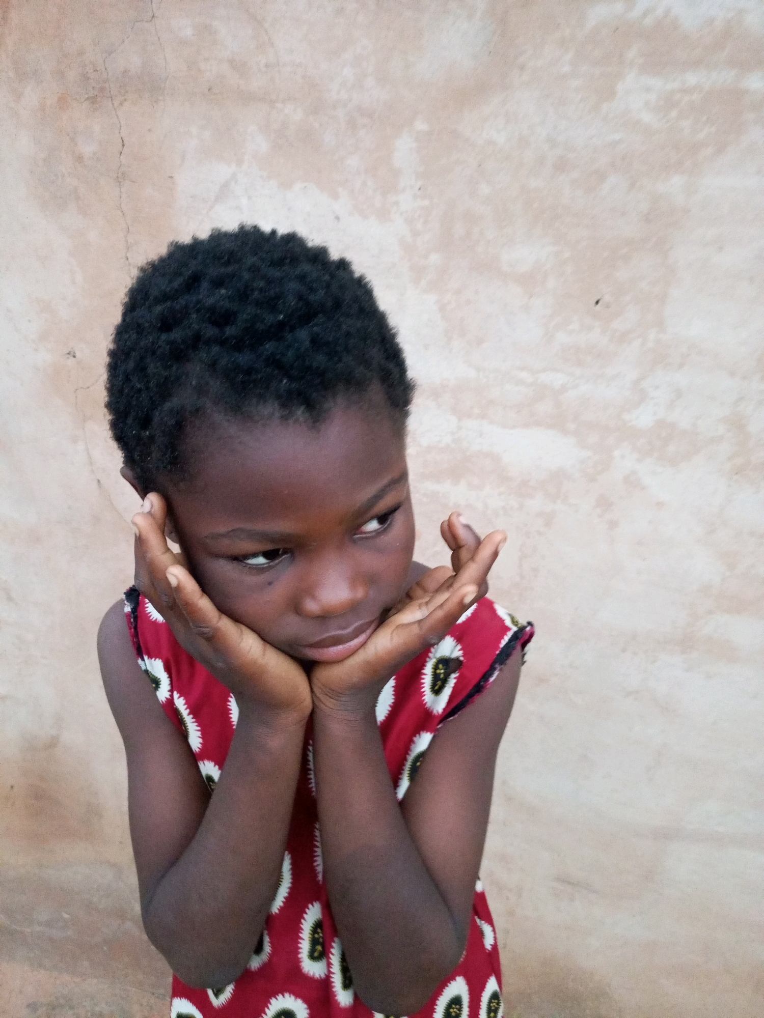 A young girl with dark skin and short, dark hair is pictured outdoors, leaning against a light-colored, textured wall. Her face is turned to her right, and she is resting her chin on her hands, with her elbows on her knees. She is wearing a red sleeveless dress with a repeating white and black circular pattern. The background wall appears to be made of a rough, possibly stucco or plaster material, with some minor cracks and variations in color. The lighting suggests it is daytime, with soft light illuminating the girl and her surroundings. The image is a close-up portrait, focusing on the girl's upper body and face. The setting appears to be a simple outdoor environment, possibly residential, in Tempure, Nigeria.