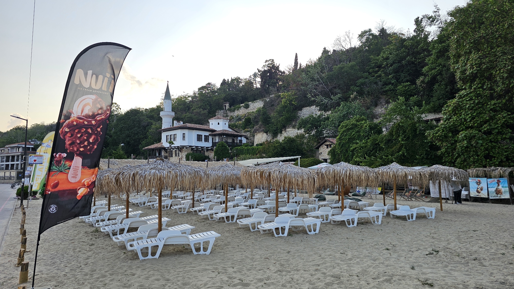 This media file depicts a tranquil beach scene in Balchik, Bulgaria. The beach is lined with numerous empty white lounge chairs and thatched umbrellas, suggesting it is either early in the morning or late in the day, or perhaps a less busy period. The sand is clean and appears well-maintained.
In the background, a picturesque white building with a distinctive minaret-like tower stands against a lush green hillside. This architectural style suggests a historical or cultural landmark. The hillside is densely covered with trees, indicating a natural and somewhat wild environment surrounding the developed beach area. A stone wall is visible on the hillside, adding to the historical feel.
To the left of the image, a tall, black flag with an advertisement for "Nuii Ice Cream Adventures" is prominently displayed. The flag features a graphic of a chocolate-covered ice cream bar with nuts. Beside it, other smaller flags and banners are visible, hinting at commercial activity on the beachfront. Further left, a street with buildings and a lamppost indicates the proximity of a town or resort.
The sky is overcast with a soft, diffused light, suggesting either dawn or dusk, and a generally calm weather condition. There are no people visible in the immediate vicinity of the lounge chairs, contributing to the peaceful and serene atmosphere of the scene. The overall impression is one of a serene coastal resort, ready to welcome visitors. Visible text includes "Nuii ICE CREAM ADVENTURES" and "Nuii".