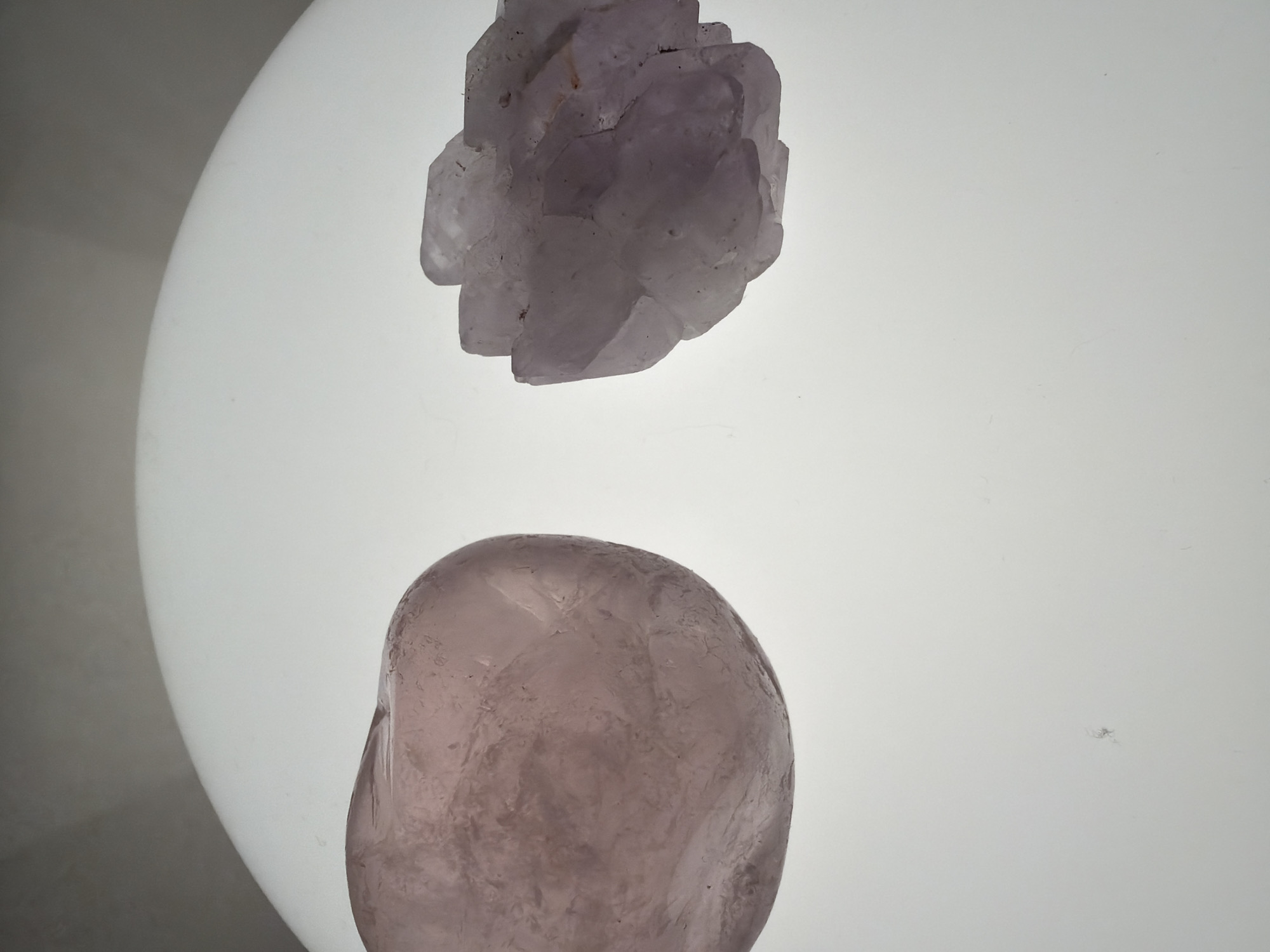 The image shows two light purple rocks on a white surface. The upper rock is larger and has a more crystalline structure, while the lower rock is smaller and smoother. The white surface appears to be illuminated from behind. There is no indication of the location being Saint Petersburg, Russia, or any other specific location. No people, actions, or interactions are depicted.