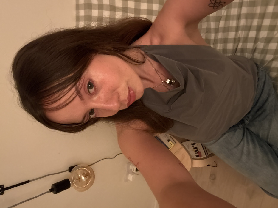 A woman with long brown hair and light skin is lying down, facing the camera. She wears a gray sleeveless top, jeans, and a silver necklace. A visible tattoo adorns her left arm. The background includes a light-colored wall with a modern lamp, books on a shelf, and a checkered blanket. The image is softly lit.