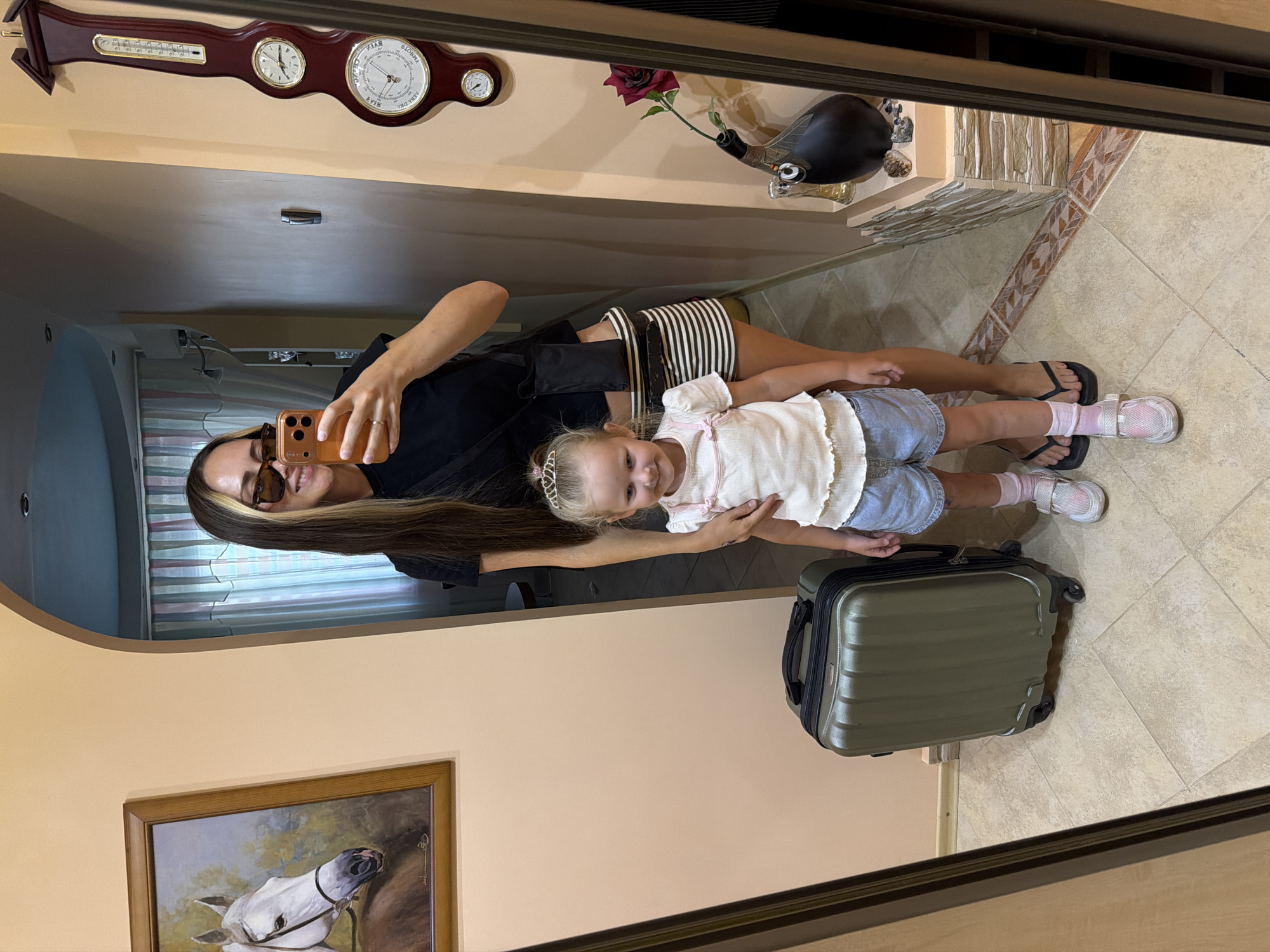 A woman and a young child are taking a mirror selfie in an indoor setting. The woman is wearing sunglasses, a black t-shirt, and a black and white striped skirt. She is holding a phone in her right hand to take the picture. The child is wearing a white top, denim shorts, and a small tiara. To the left of the frame is a large, dark green suitcase. In the background, there is a painting of a white horse on the wall, and a large barometer on the right wall. The floor is tiled. The setting appears to be a hallway or entryway, possibly in a residential building. There is no specific information in the image to suggest a location in Kamyshin, Russia.