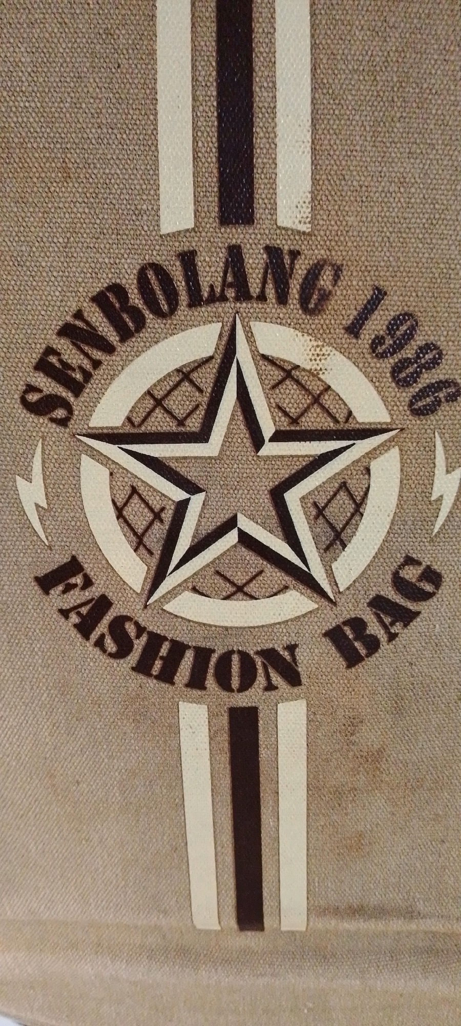 The image shows a close-up view of a textured fabric, likely a bag, with a graphic design. The design features a large star in the center, outlined in dark brown and light beige, with a grid pattern inside. Arched text above the star reads "SENBOLANG 1986" in dark brown letters. Arched text below the star reads "FASHION BAG" in dark brown letters. Two vertical stripes, one dark brown and two light beige, run down the center of the fabric, partially covered by the star design. Small lightning bolt shapes are visible on either side of the star. The fabric appears to be a neutral, light brown or tan color with a visible weave. The image does not contain any information or visual cues that would allow for its location to be identified in Jalingo, Nigeria, or any other specific urban environment.