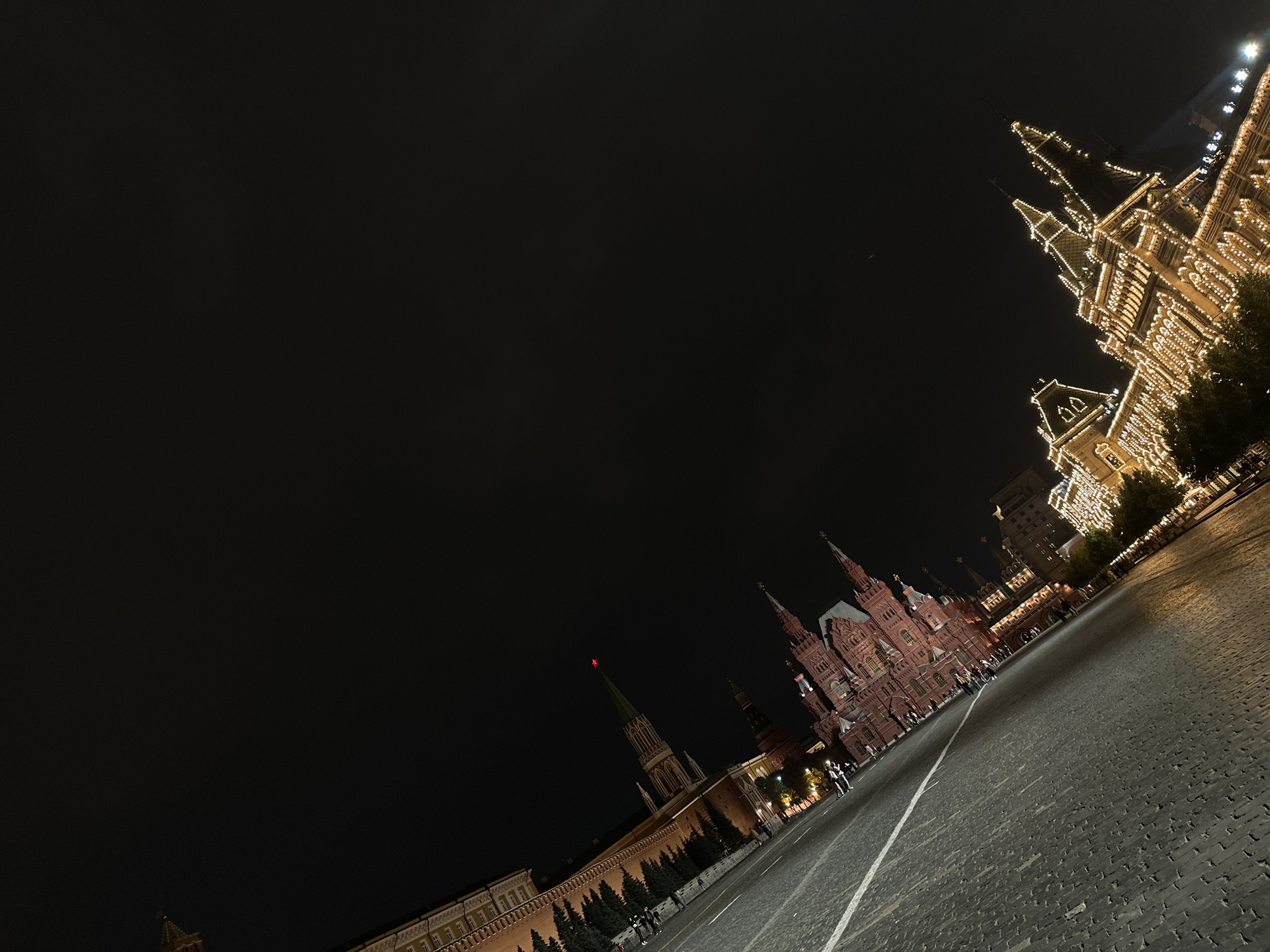 The image depicts Red Square in Moscow at night. The scene is dominated by a dark sky, with artificial lighting illuminating several historic buildings. On the right side of the frame, a grand building, likely the GUM department store, is adorned with numerous lights, creating a festive and striking appearance with its spires and intricate architectural details. In the mid-ground, the distinctive red facade of the State Historical Museum is visible, also illuminated. A red star tops a tall spire in the distance, a recognizable symbol of Russia.
The ground of Red Square, paved with cobblestones, stretches towards the buildings, with white lines marking pathways or divisions. Several people are present, scattered across the square, appearing as small figures against the monumental architecture. They seem to be strolling or observing the illuminated buildings, suggesting a leisurely evening activity. The overall mood is one of grandeur and perhaps awe, amplified by the dramatic night lighting and the historical significance of the location. The time of day is clearly night, and the weather appears clear, allowing for good visibility despite the darkness. There is no visible text in the image.