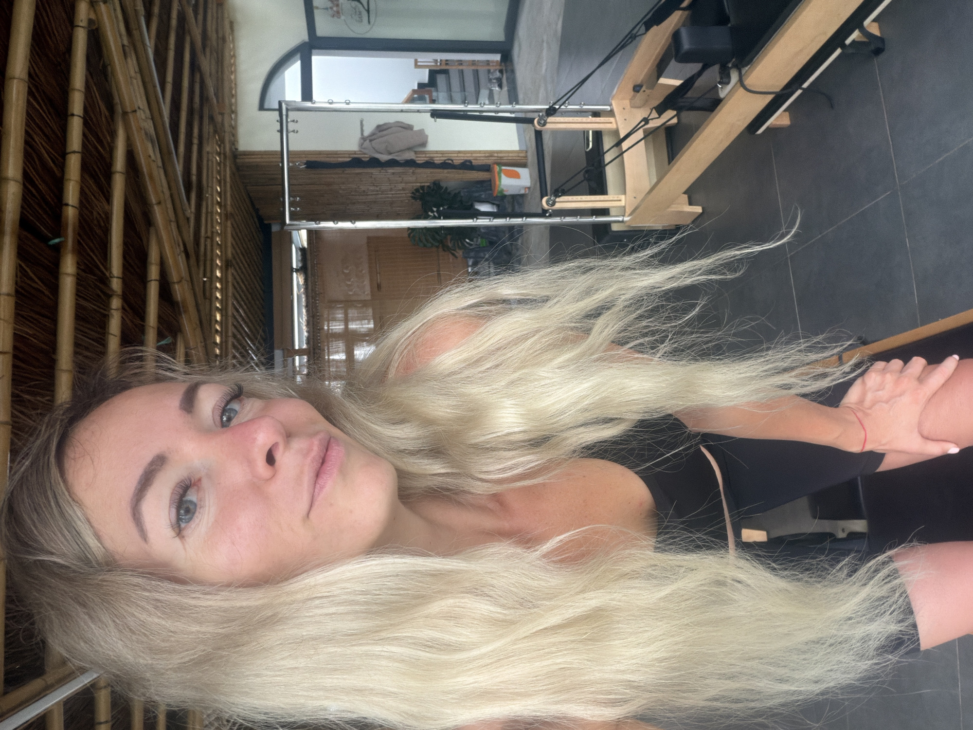 An over-the-shoulder, medium selfie captures a young Caucasian woman with long, blonde hair, a black athletic top, and black leggings. She is positioned in what appears to be a Pilates studio with a wooden ceiling structure and bamboo accents. In the background, a Pilates reformer machine is visible, along with a mirror and a staircase. The woman is leaning forward, resting her hands on her knees, and looking directly at the camera with a slight smile. Her right wrist is adorned with a thin red string bracelet. The lighting suggests it is daytime. The overall setting appears to be an indoor fitness or wellness space, likely in a tropical or semi-tropical climate given the architectural style and natural materials.