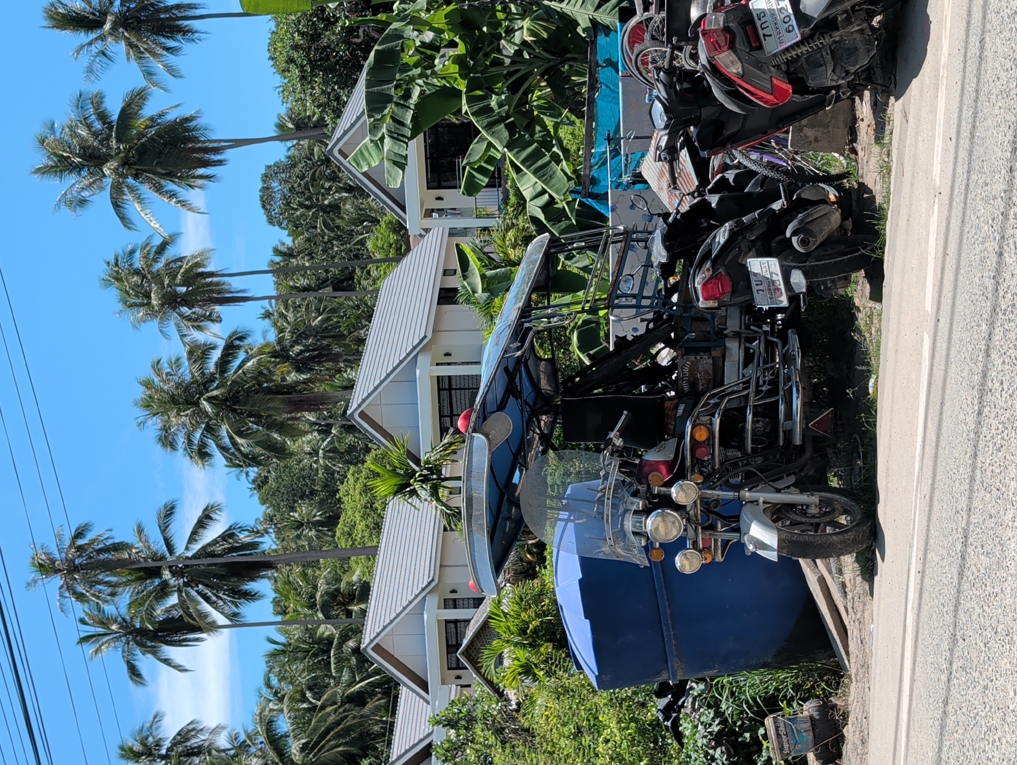 The image shows a street scene in ตำบลเกาะพะงัน, Thailand. Palm trees are visible against a clear blue sky, with several houses and lush vegetation in the background. Parked along the roadside are a variety of motorcycles, including a blue tuk-tuk with a white trim and various other motorbikes, one of which is red and black. The street is made of asphalt and shows signs of wear. There are no people visible in the image.