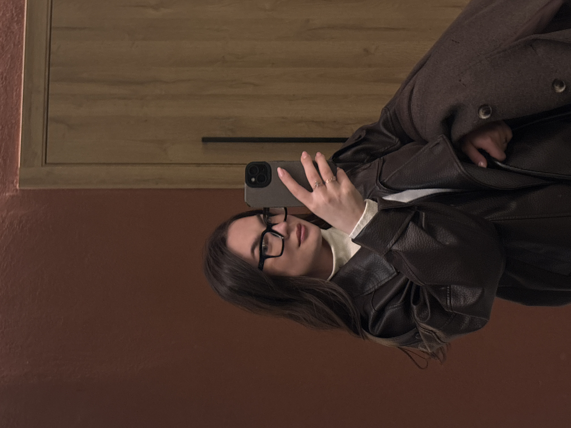 A woman with long, dark hair and glasses is taking a selfie with her phone. She is wearing a dark brown leather jacket over a white top. Part of a grey wool coat is also visible. The background consists of a brown wall and a wooden cabinet. The location is not discernible from the image.