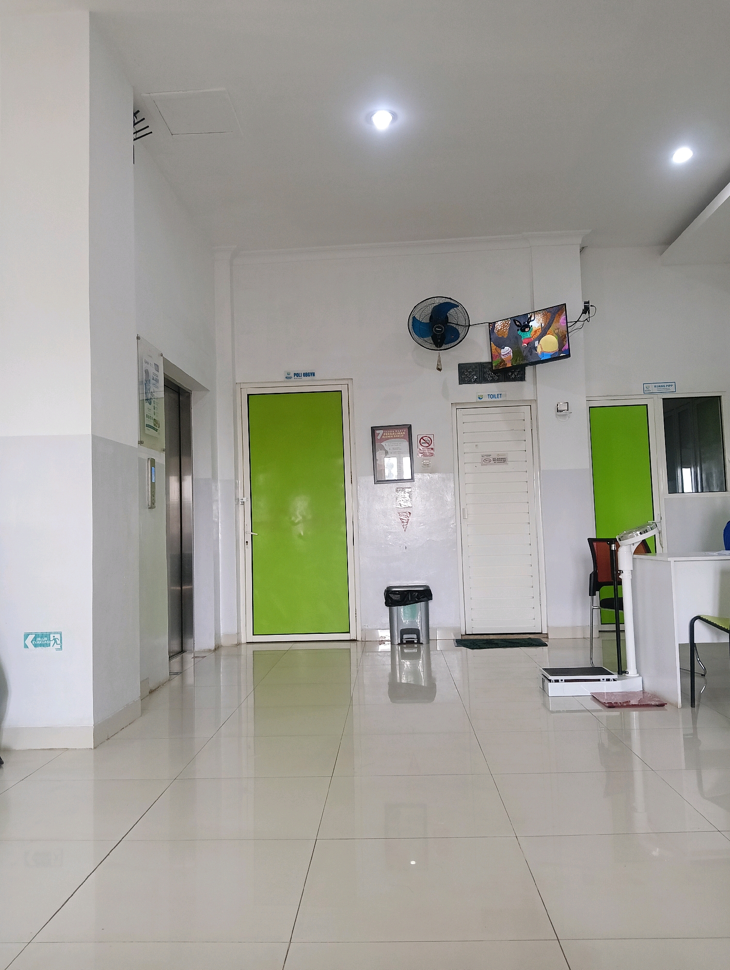 The image depicts an indoor hallway scene, possibly within a medical facility, in Purwareja, Indonesia. The hallway has a white, tiled floor that reflects the overhead lighting. On the left side, there is a silver elevator with a control panel on the wall next to it. An "Evacuation Route" sign with a green running figure is visible on the lower left wall.
Further down the hallway, on the left, is a bright green door labeled "POLI OBGYN" in blue text. To the right of this is a white door labeled "TOILET" in blue text. Above the toilet door, a television is mounted on the wall, displaying a cartoon image. A blue oscillating fan is also mounted on the wall above the television. A black trash can is positioned in front of the toilet door. To the right of the toilet door, a chair is visible.
Further to the right, there is another bright green door. In the foreground on the right side of the hallway, a white medical scale is situated on the floor, with a pink mat in front of it. A white counter with a dark object on it is also present on the right side. The walls are white, with some architectural trim visible near the ceiling. The overall impression is that of a clean, functional, and brightly lit corridor.