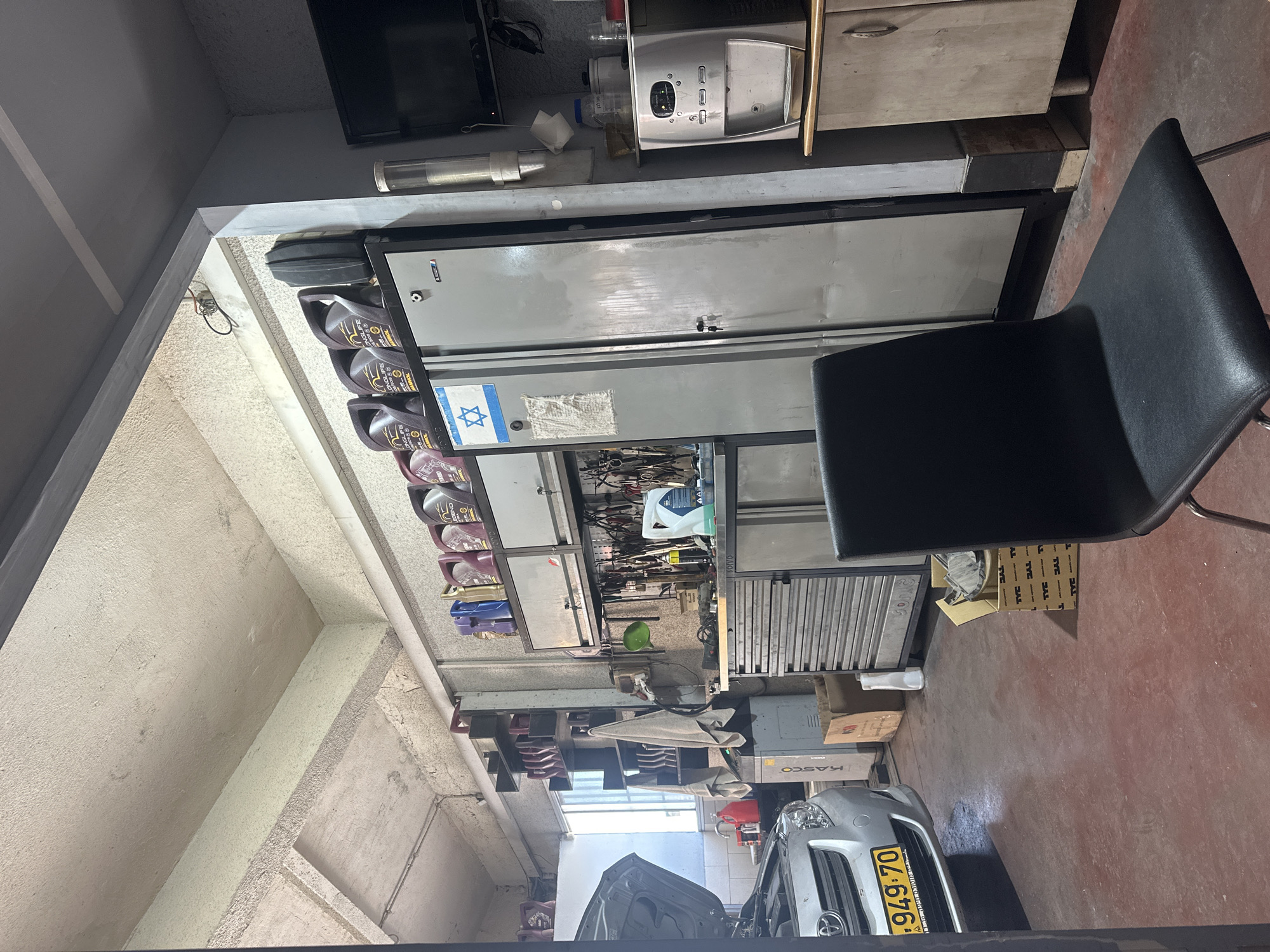The image depicts the interior of a workshop, likely a car repair shop, located in Haifa, Israel. The workshop is filled with various tools and equipment, suggesting a functional space for vehicle maintenance. In the foreground, a black chair with metal legs is positioned centrally. To the left, a silver car with its hood open is parked, its license plate reading "949 70". The floor is a reddish-brown color, indicative of a workshop environment.
Dominating the background is a tall, grey metal cabinet with multiple drawers and doors, a common storage unit in such settings. On top of this cabinet, several large jugs of oil are neatly arranged, with some displaying "LONGLIFE" and "SYNTHETIC" labels. A blue and white Israeli flag sticker is affixed to one of the cabinet doors. To the right of the cabinet, a water dispenser is visible, alongside a wooden cabinet unit. Tools are neatly organized on a pegboard mounted on the wall behind the cabinet. Shelving units are also present, holding more containers and supplies. The ceiling appears to be a textured concrete structure, with exposed conduits and pipes. The overall impression is one of a well-organized, operational workshop.
