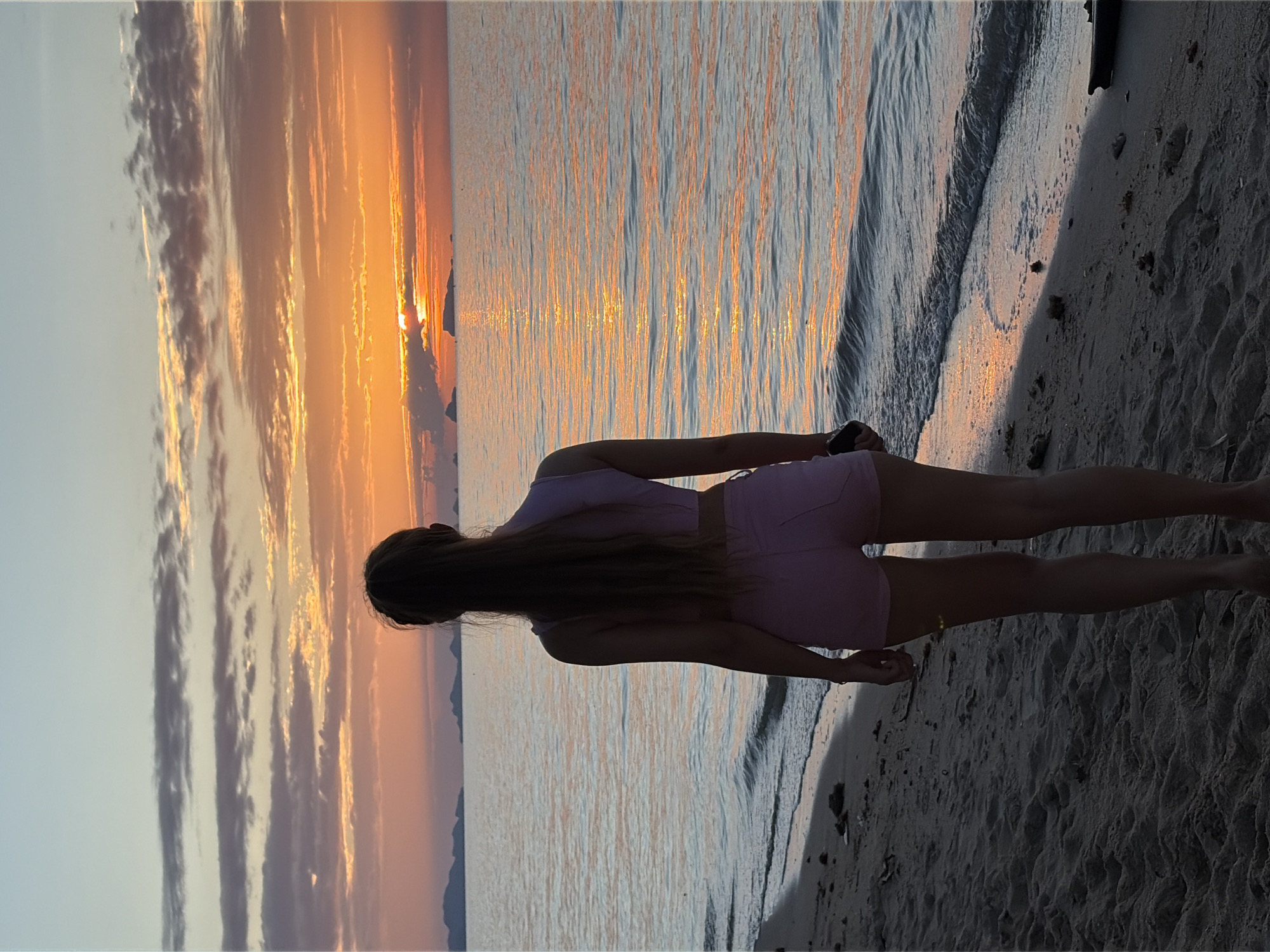A rear view of a person standing on a sandy beach facing a sunset over the ocean. The person is wearing a light purple crop top and pink shorts. The sun is low on the horizon, casting an orange glow on the water and clouds. In the distance, several islands are visible. The beach is covered in sand and some debris. The water is calm with gentle waves lapping at the shore. The sky is a mix of blue and orange with scattered clouds. The location is Baan Thong Po, Thailand.