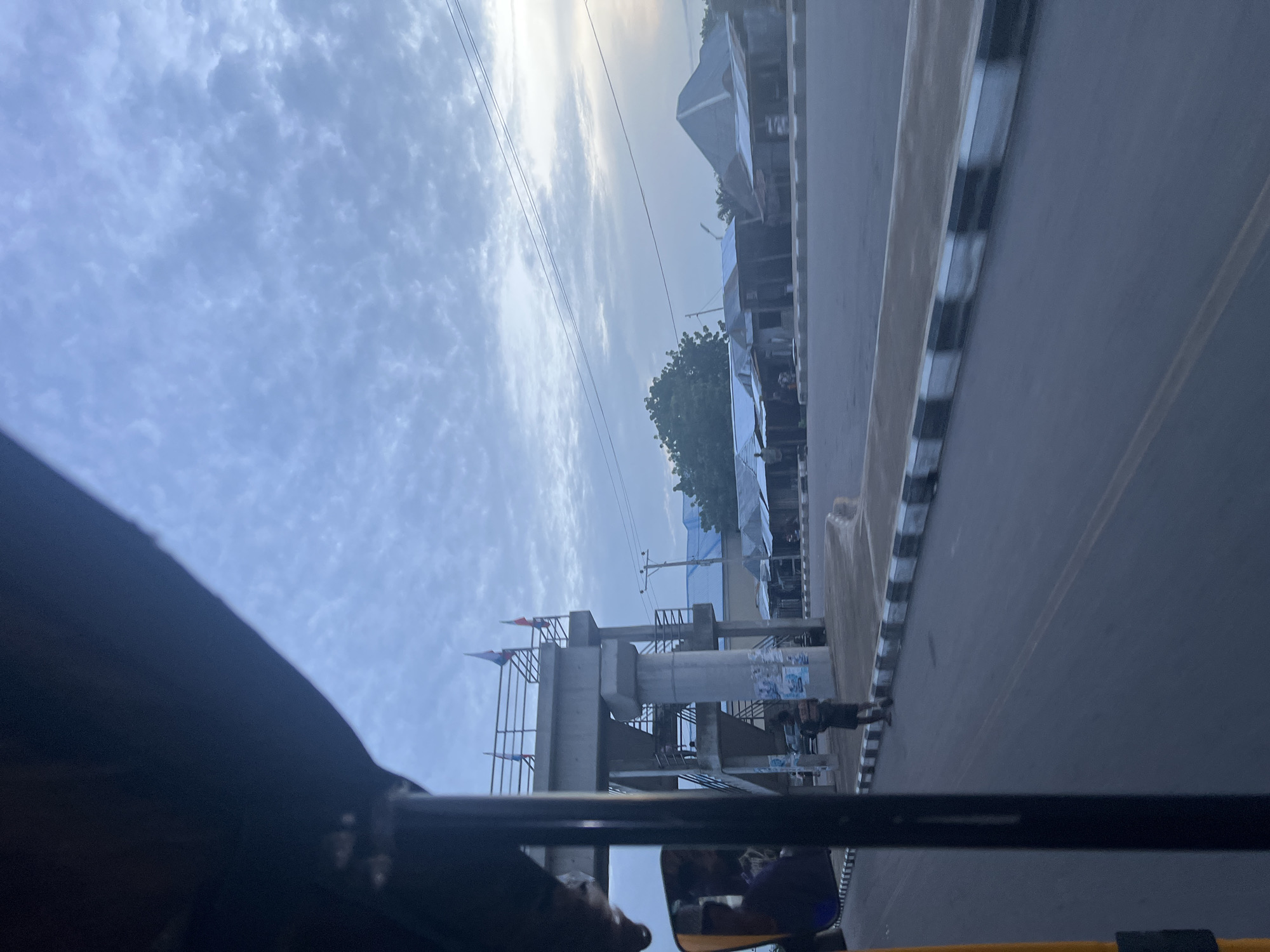 The image captures a view from inside a vehicle, likely a tricycle, looking out towards a street scene in Jalingo, Nigeria. In the foreground, the dark interior of the vehicle is visible, along with a yellow component and a side mirror reflecting a person's head. The street ahead features a pedestrian overpass, a concrete structure with stairs and a railing, adorned with flags. A person stands on the sidewalk near the overpass. Along the side of the road are several structures with corrugated metal roofs, appearing to be shops or small buildings. The sky is overcast with scattered clouds. The environment suggests an urban or semi-urban setting with infrastructure and commercial activity.
