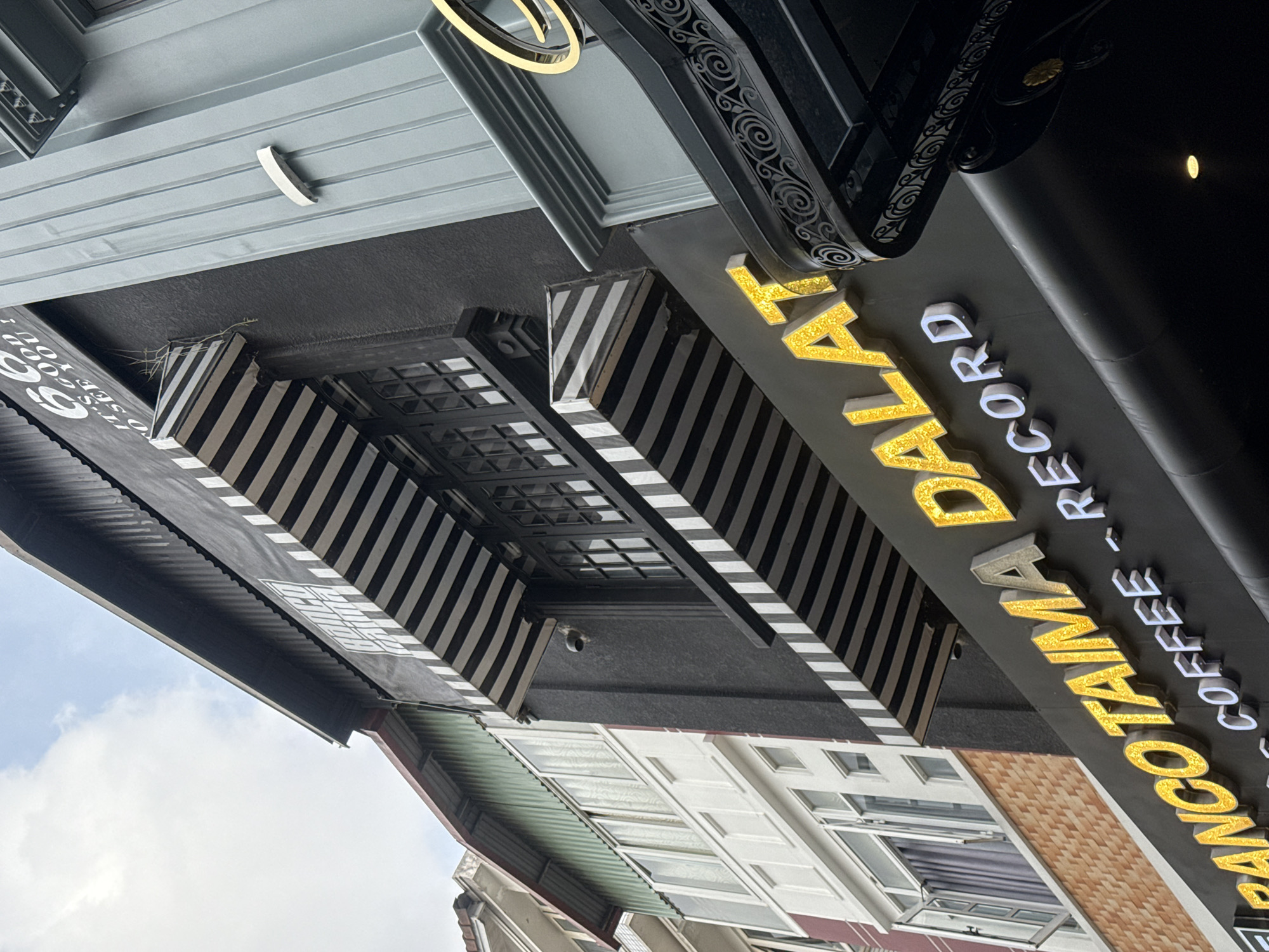 An exterior, low-angle, close-up shot captures the facade of a building in Da Lat, Vietnam. The building features a dark gray painted exterior with black and white striped awnings above windows. The building's signage is prominent, with large, yellow, glitter-filled letters spelling out "BANCO TAIMA DALAT". Below this, smaller white letters read "COFFEE - RECORD". To the right, a decorative gray column is visible. The sky above is cloudy and pale. The architecture suggests a modern establishment, possibly a cafe or shop, situated within the urban landscape of Da Lat.