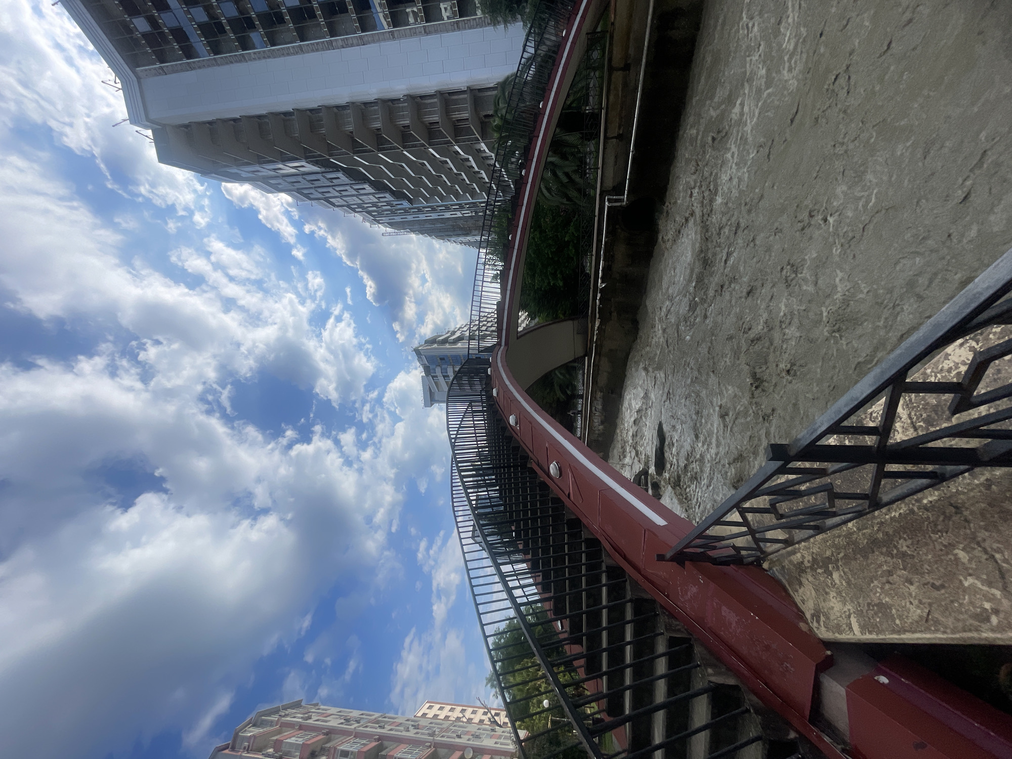 The image depicts a view from a red pedestrian bridge overlooking a fast-flowing, murky river. The bridge has a dark metal railing and stairs on the left side. In the background, there are several tall, multi-story buildings, some of which appear to be under construction. The sky is blue with scattered white clouds. The scene is likely in Sochi, Russia, given the architectural style of the buildings and the presence of palm trees visible amongst the foliage. No people are visible in the image.