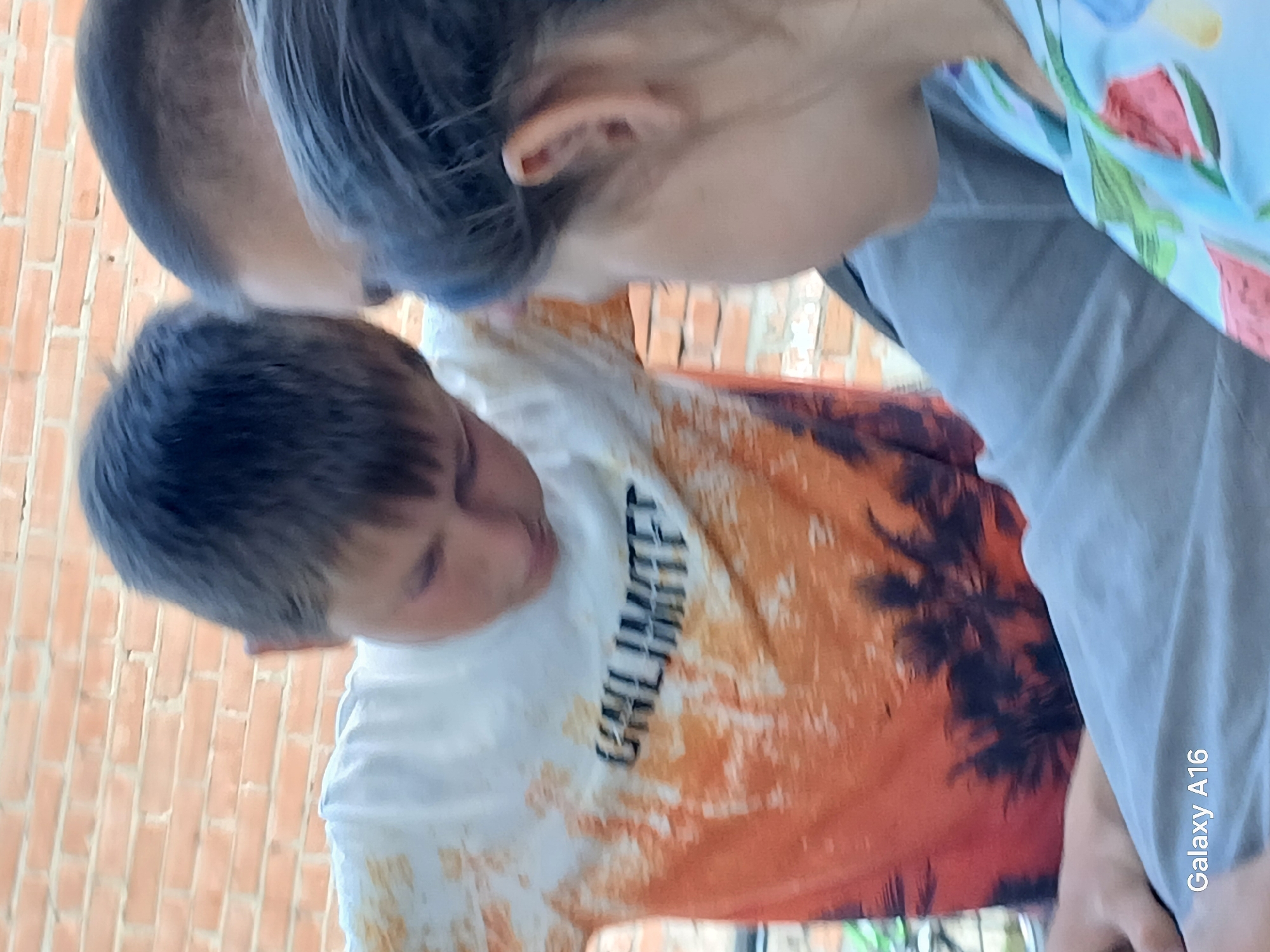 The image shows three people, appearing to be children, in an outdoor setting with a brick wall in the background. The central figure, a boy wearing a white t-shirt with an orange and black palm tree design, is looking down. To his left is another boy with dark hair. To his right is a person whose face is partially visible, with dark hair and an ear clearly showing. This person is wearing a blue shirt with a floral or fruit pattern. The lower right corner of the image displays "Galaxy A16," indicating the device used to capture the photograph. The environment suggests a casual gathering. There is no specific information in the image to pinpoint a location in the city of Ustyvytsia, Ukraine.