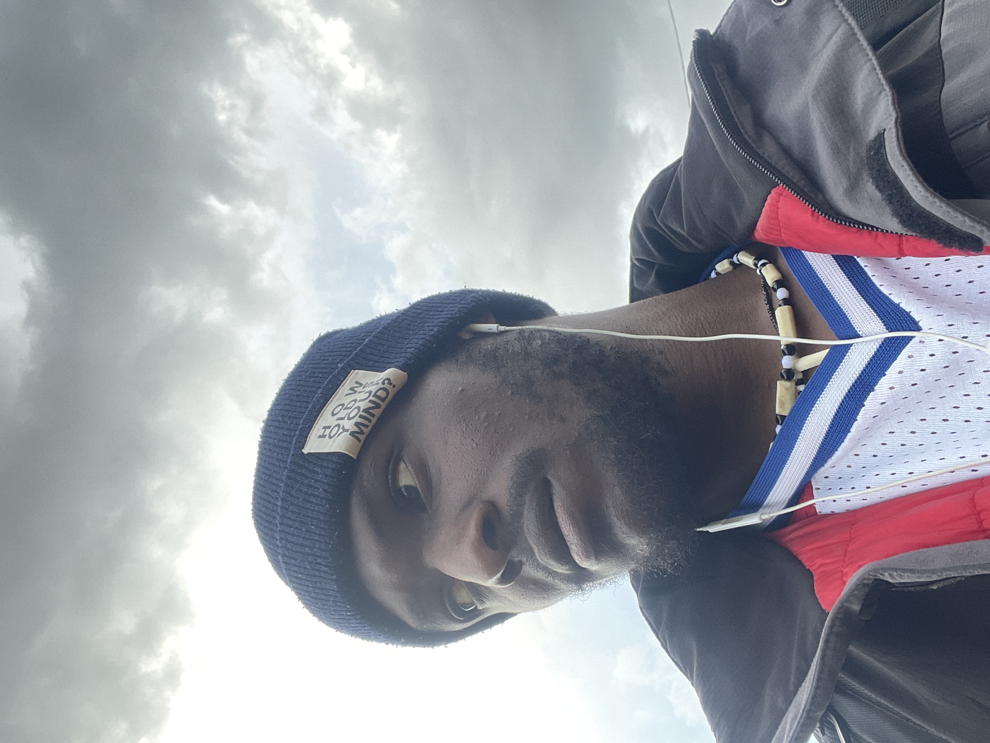 A man wearing a blue beanie with a patch that reads "HOW YOU MIND?" is looking to the left. He has earbuds in and is wearing a black jacket over a red and white mesh jersey with blue and white trim. He is also wearing a necklace made of wooden beads and black beads. The background is a cloudy sky, suggesting an outdoor setting, possibly in Jos, Nigeria, given the context provided.