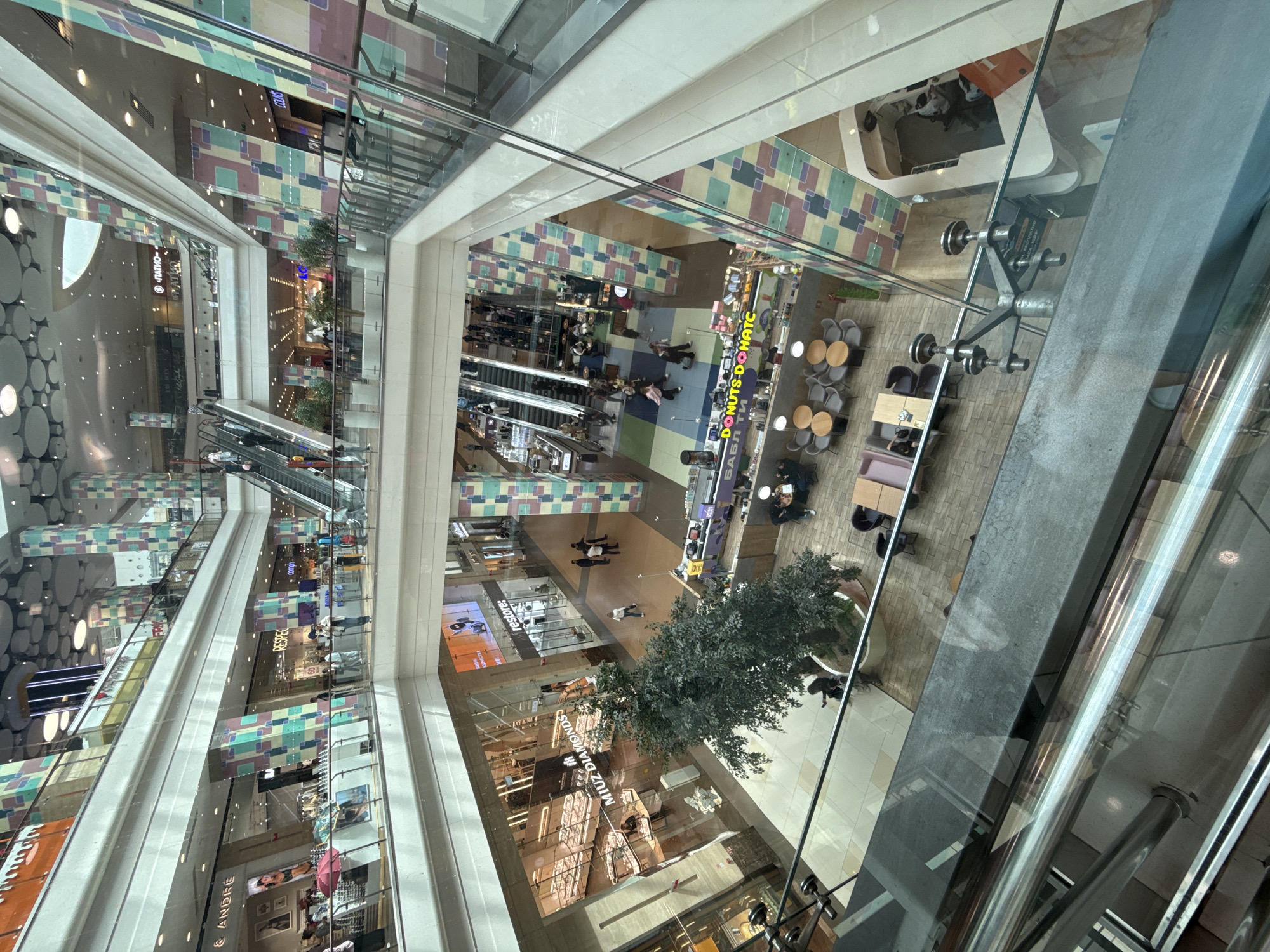 The image shows the interior of a multi-level shopping mall in Moscow, Russia. The mall features escalators, shops, and food establishments. In the foreground, a glass railing overlooks the lower levels. People are seen walking, shopping, and sitting at tables in the mall. The architecture of the mall is modern, with columns decorated in a colorful, geometric pattern. A large potted tree is visible in the center of the image. A food stall labeled "DONUTS DONATC" is present on one of the lower levels. The overall impression is a busy and lively commercial space.