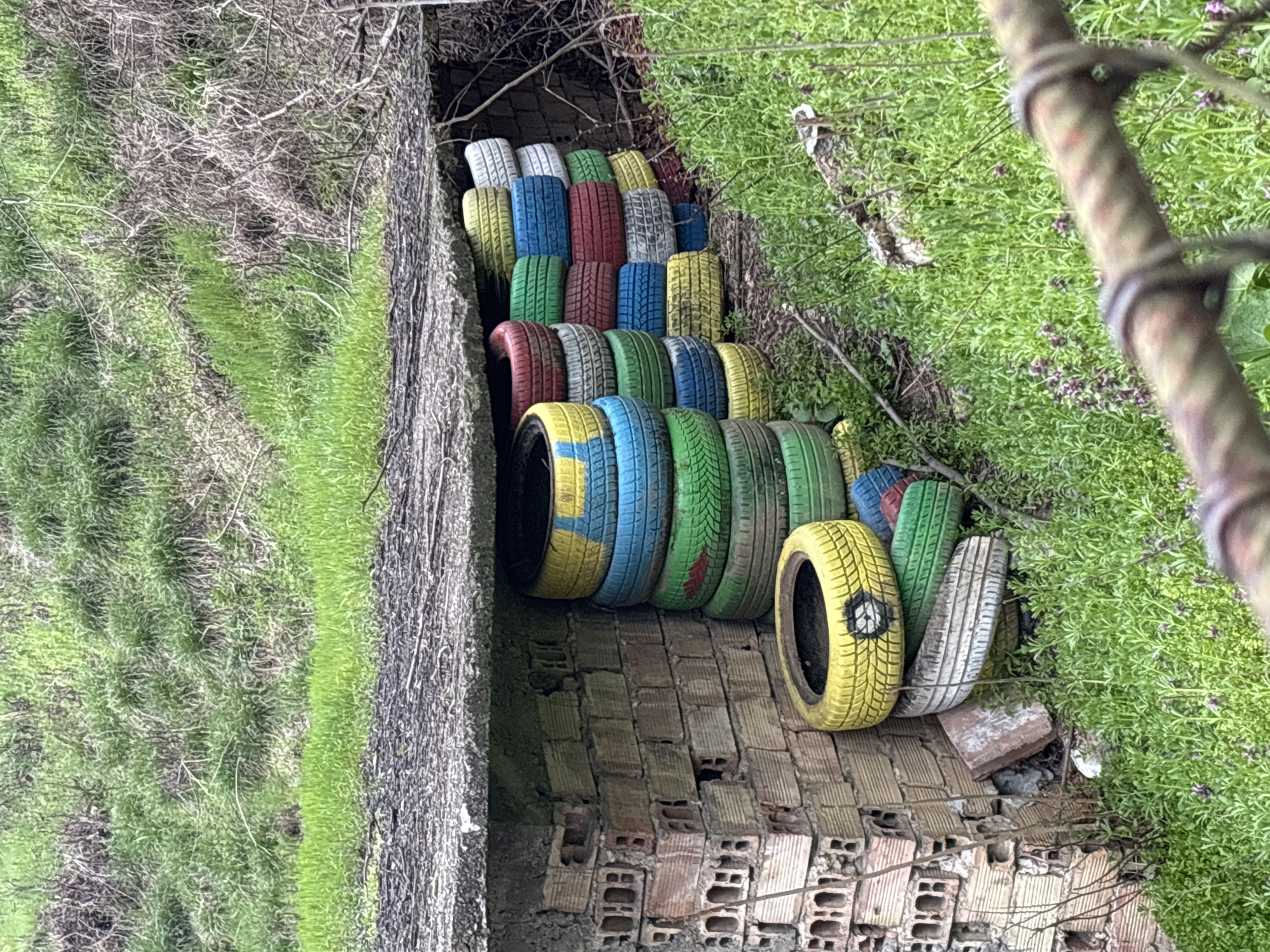 A partially collapsed brick structure serves as a base for a collection of painted tires. The tires ascend in a stepped fashion, forming a makeshift staircase or decorative feature. The tires are painted in various colors, including yellow, blue, green, red, and white. Lush green vegetation flanks both sides of the structure and the tire arrangement. The structure is located on the side of the road going up to Tsarevets Fortress in Veliko Tarnovo, Bulgaria.