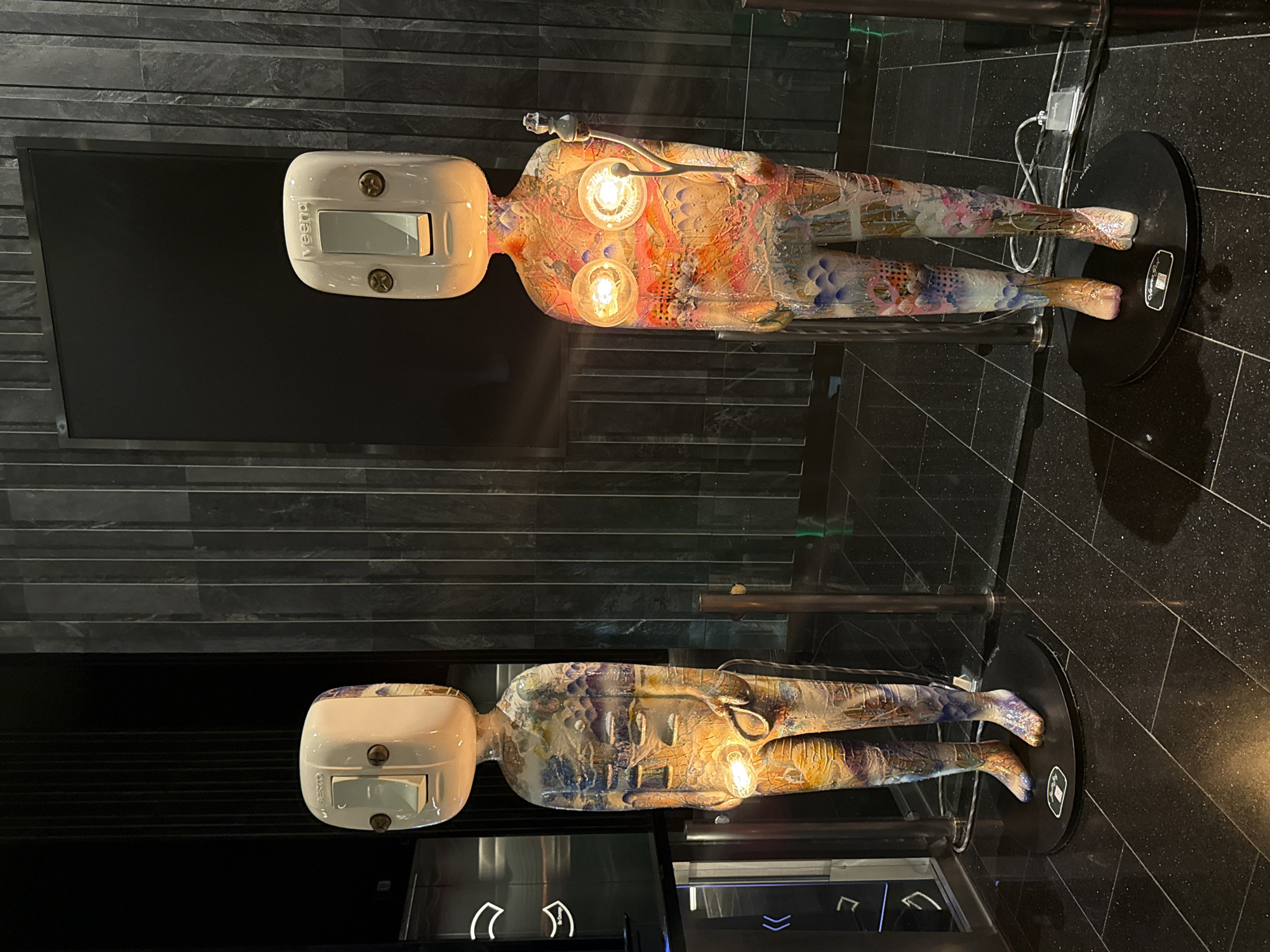 The image depicts a modern art installation in Bangkok, Thailand. Two abstract human-like figures lie horizontally on black, circular stands, their bodies painted with intricate patterns and illuminated by internal lights. The figures have blocky, white heads with rectangular features. The background consists of a dark, horizontally-patterned wall and a shiny, black tiled floor. A large, dark rectangle hangs on the wall behind the top figure. The figures appear to be displayed in a gallery or public space with a reflective surface beneath them, creating a mirrored effect.