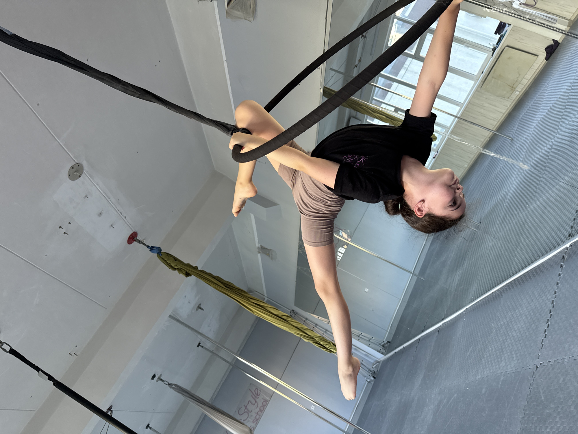 The media file shows a young female performing aerial silks in an indoor studio. She is upside down, suspended by black silks, with her body in a contorted pose. Her legs are extended, and her arms are wrapped around the silks. She is wearing a black t-shirt and light brown shorts. The studio appears to have a high ceiling with exposed beams and lighting fixtures. There are also mirrors on the walls and padded flooring. A green aerial hammock is visible in the background, along with other rigging equipment. The scene is well-lit, suggesting it is daytime. The overall atmosphere seems to be one of athletic training or performance. A partial view of a sign with the text "Style School" is visible on the floor. The location is specified as Dnipro, Ukraine.