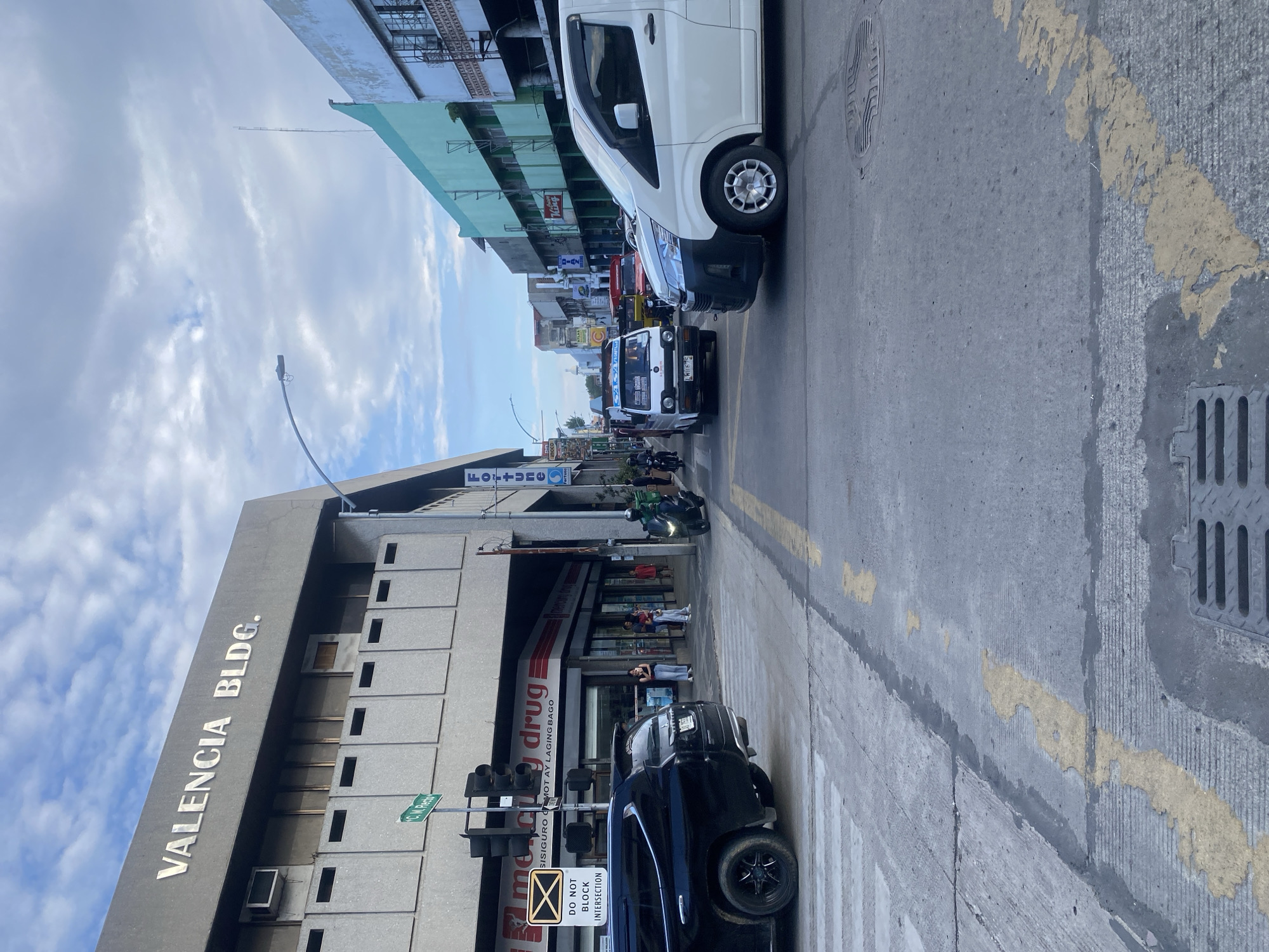 The image captures a street scene in Davao City, Philippines, likely near the Valencia Building. A black SUV is positioned in the foreground, facing left at an intersection. To its right, a white van is also stopped at the intersection. Behind the vehicles, a Mercury Drug store is visible with people standing on the sidewalk. The street is paved with asphalt, marked with yellow lines and a pedestrian crossing. Traffic lights are visible, indicating an active intersection. The buildings lining the street show a mix of architectural styles, with some appearing older and others more modern. The sky is partly cloudy, suggesting daytime. A street sign labeled "C. M. Recto" is mounted on a traffic light pole, confirming the location is indeed along C. M. Recto Street in Davao City.