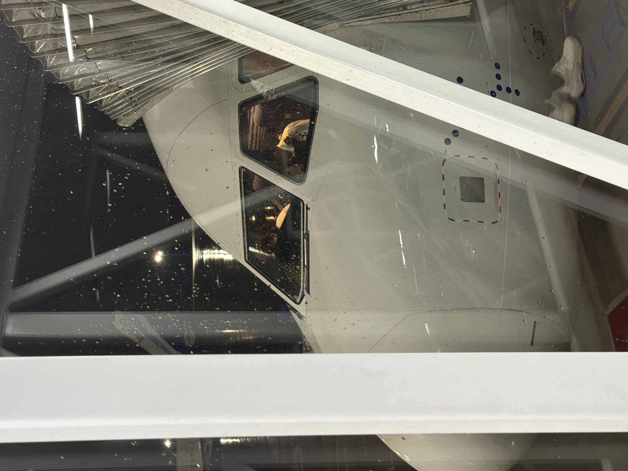 The image shows the cockpit of an airplane viewed from the exterior through a jet bridge. The airplane is white with visible water droplets on its surface, suggesting it might be raining or the image was taken in a humid environment. The jet bridge is a metallic, accordion-like structure that connects the terminal to the aircraft. Inside the cockpit, two pilots can be seen, their faces partially obscured. The pilot on the left is wearing a dark uniform, and the pilot on the right appears to be wearing a lighter-colored shirt. The cockpit windows are also covered in water droplets, distorting the view of the interior slightly. In the background, through the cockpit windows, there are lights and reflections indicating an airport environment at night. The image also shows a portion of the airplane's fuselage with some markings, including a square with a dashed red outline and a series of blue circular markings. A pair of white sneakers is visible on the ground near the base of the aircraft. The scene appears to be at Rajiv Gandhi International Airport in Hyderabad, India, given the Indigo Airlines logo visible on the aircraft.