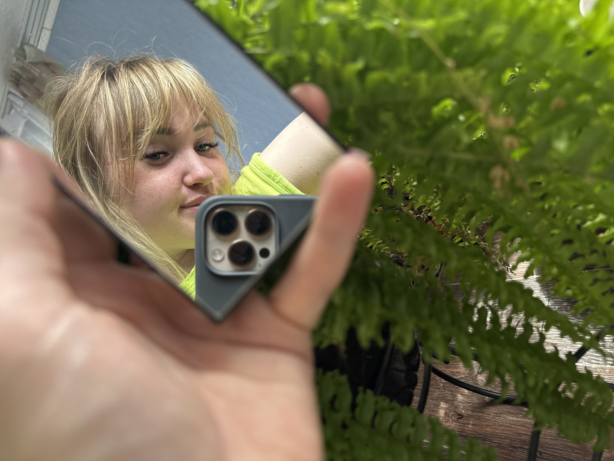 A woman with light blonde hair and bangs is reflected in a small, tilted mirror held in her left hand. She is wearing a neon yellow top. The mirror also reflects a portion of a bright blue sky. The hand holding the mirror is blurred. Behind the mirror and hand, on the right side of the frame, is a lush green fern plant. A black metal structure is visible beneath the fern. It is not possible to determine the exact location in the city of Zavod Mikhailovskii, Russia based solely on this image.