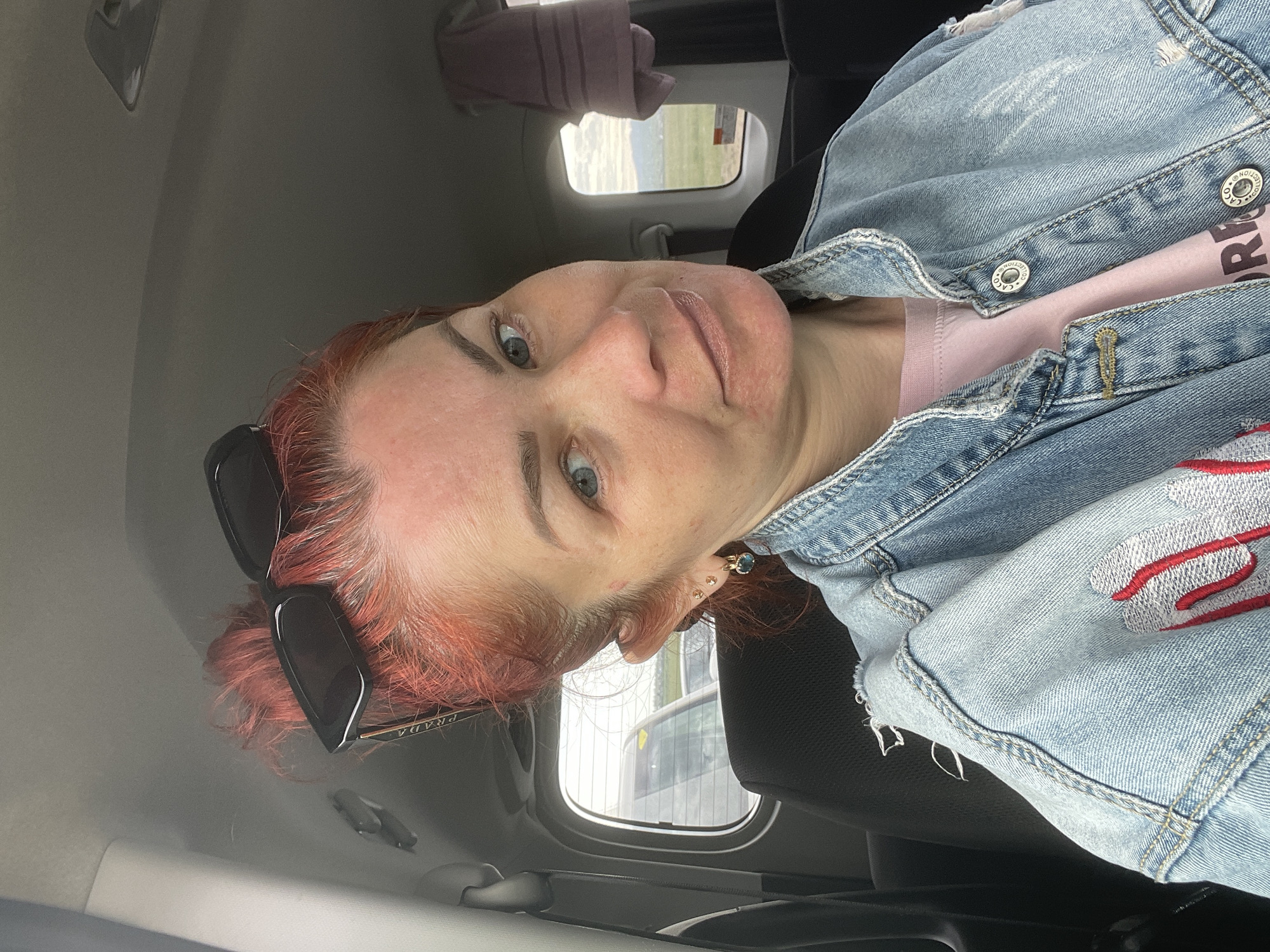 A woman with red hair is sitting in the passenger seat of a car. She is wearing a light pink t-shirt and a light blue denim jacket with red and white embroidery on the sleeve. Her sunglasses are on top of her head. The car interior is visible, including the dark seats and the car's ceiling. Through the car window, a slightly overcast sky and some distant greenery can be seen. The image appears to be taken indoors, possibly in a parking lot or on a road. The woman's expression is neutral.