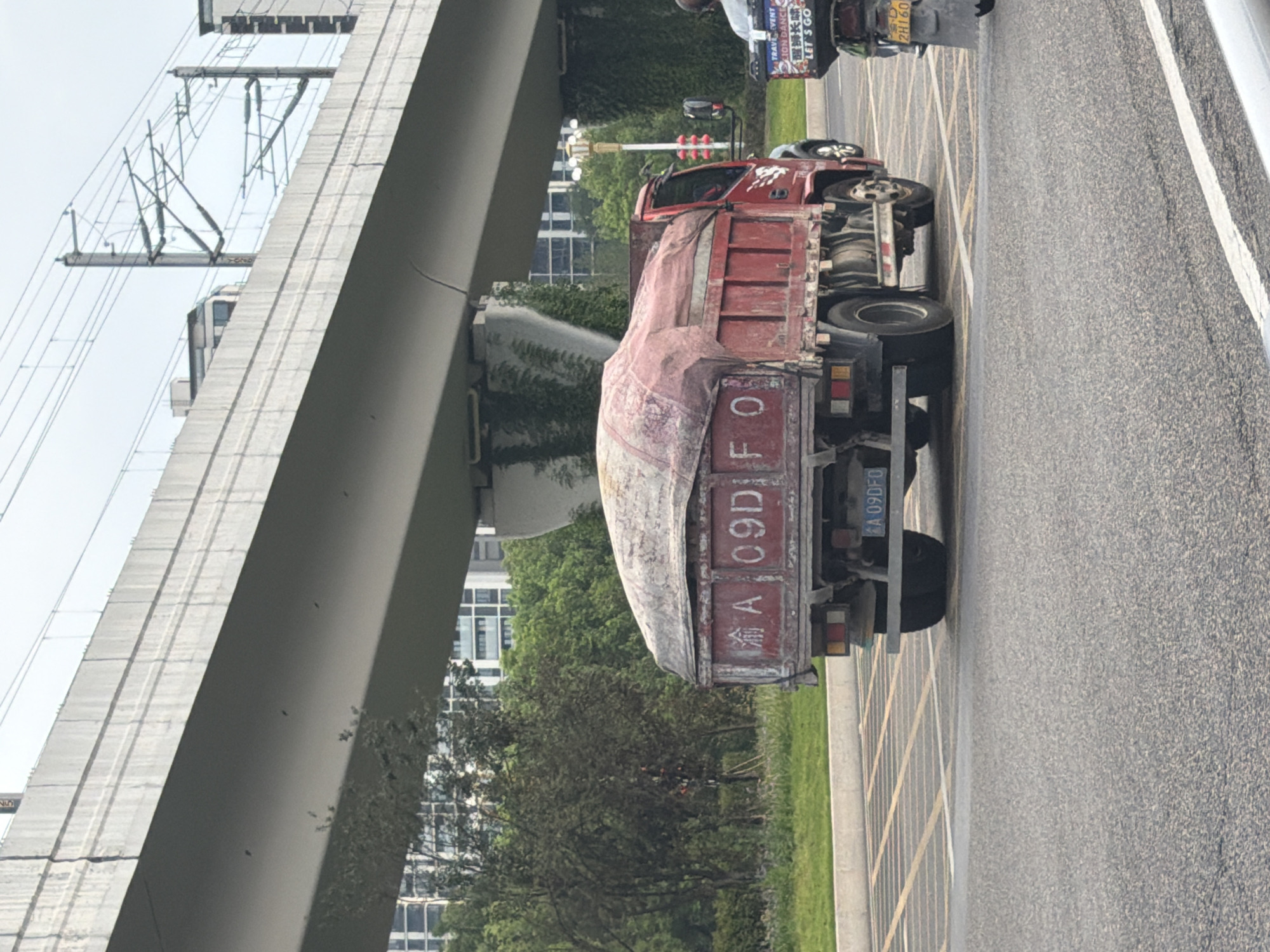 The image shows a red dump truck driving under an elevated overpass. The truck has a tarp covering its load, and a license plate that reads "渝A 09DFO". In the background, there are tall buildings and trees. To the right of the truck, a scooter is visible with a passenger. The road appears to be in an urban area. The license plate starting with "渝A" indicates that the vehicle is registered in Chongqing, China.