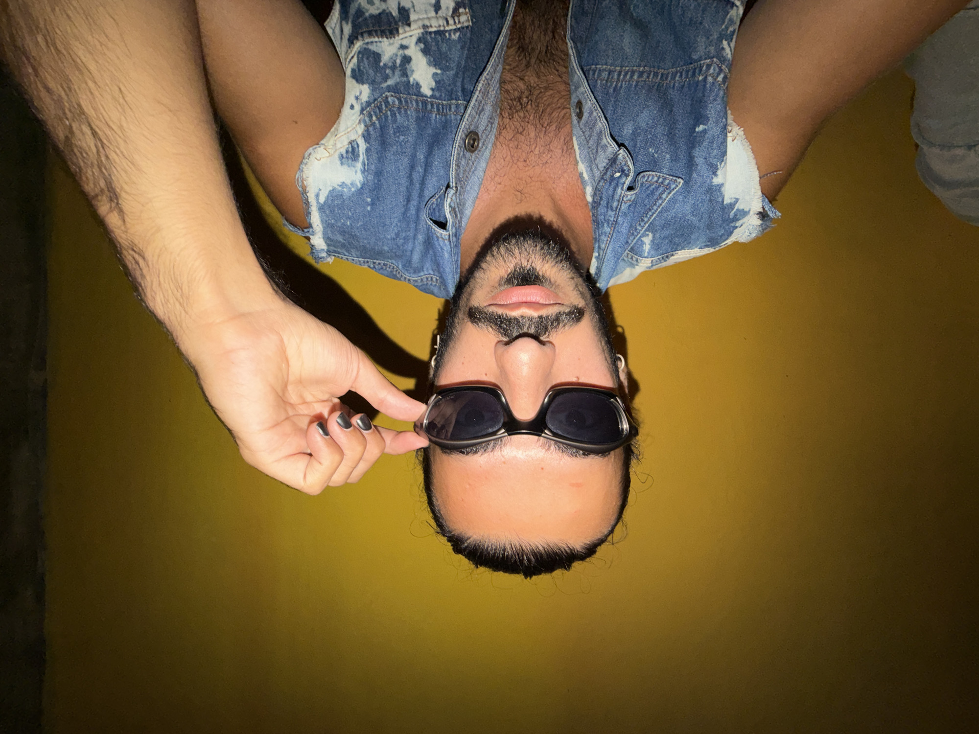 A man with dark hair pulled back and a goatee is posing for a photo against a yellow wall. He is wearing sunglasses and a distressed denim vest. His right hand is raised, adjusting his sunglasses. The image is a close-up shot, focusing on his face and upper body. The lighting is dramatic, with a strong light source from the front casting shadows. The man appears to be in an indoor setting, possibly a room or studio. The background is a solid yellow color, with some subtle variations in shade. There is a hint of a dark, vertical element on the right side of the frame, suggesting a wall or doorway. The overall mood of the image is casual and confident. No specific details about North Kuta, Indonesia are visible in the image.