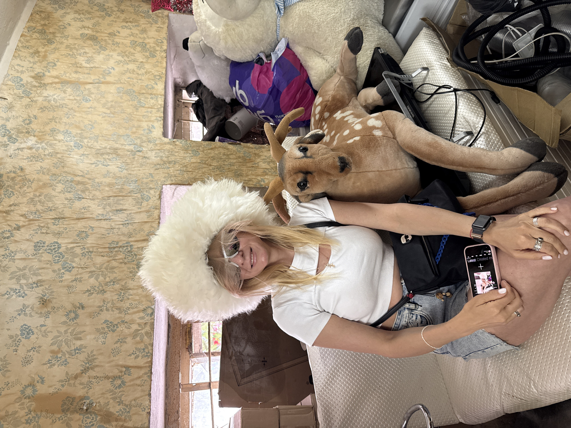 A forensic, matter-of-fact description of the scene in the city of Unknown, Unknown:
A young adult female is seated next to a large plush deer toy in a cluttered indoor environment. The female has blonde hair, wears a large white fluffy hat and white-framed mirrored sunglasses. She is dressed in a white ribbed crop top, light blue denim shorts, and a black crossbody bag with a blue chain strap. Her left hand holds a pink smartphone displaying a media player interface, while her right hand, adorned with multiple rings, rests on her leg. A pearl-like bracelet is visible on her left wrist, and an Apple Watch is on her right. She is smiling directly at the viewer.
The plush deer, brown with white spots and small antlers, lies on a black object next to the female, facing the camera. Both are positioned on a light-colored, textured upholstered surface, possibly a couch or bench.
The background features old, distressed wallpaper with a faded beige-yellow base and light blue floral patterns. To the left, a window with visible wooden framing and an outside view of greenery is present, along with stacked cardboard boxes, one of which shows partial text. To the right, a large pile of assorted items includes a big white plush polar bear with a blue and white striped scarf, another large white plush toy, and a purple plastic bag. Below these items, a white pillow, various cables, and cardboard debris are visible on the floor. The overall environment appears to be an informal, cluttered space, possibly a storage room, illuminated by natural light.