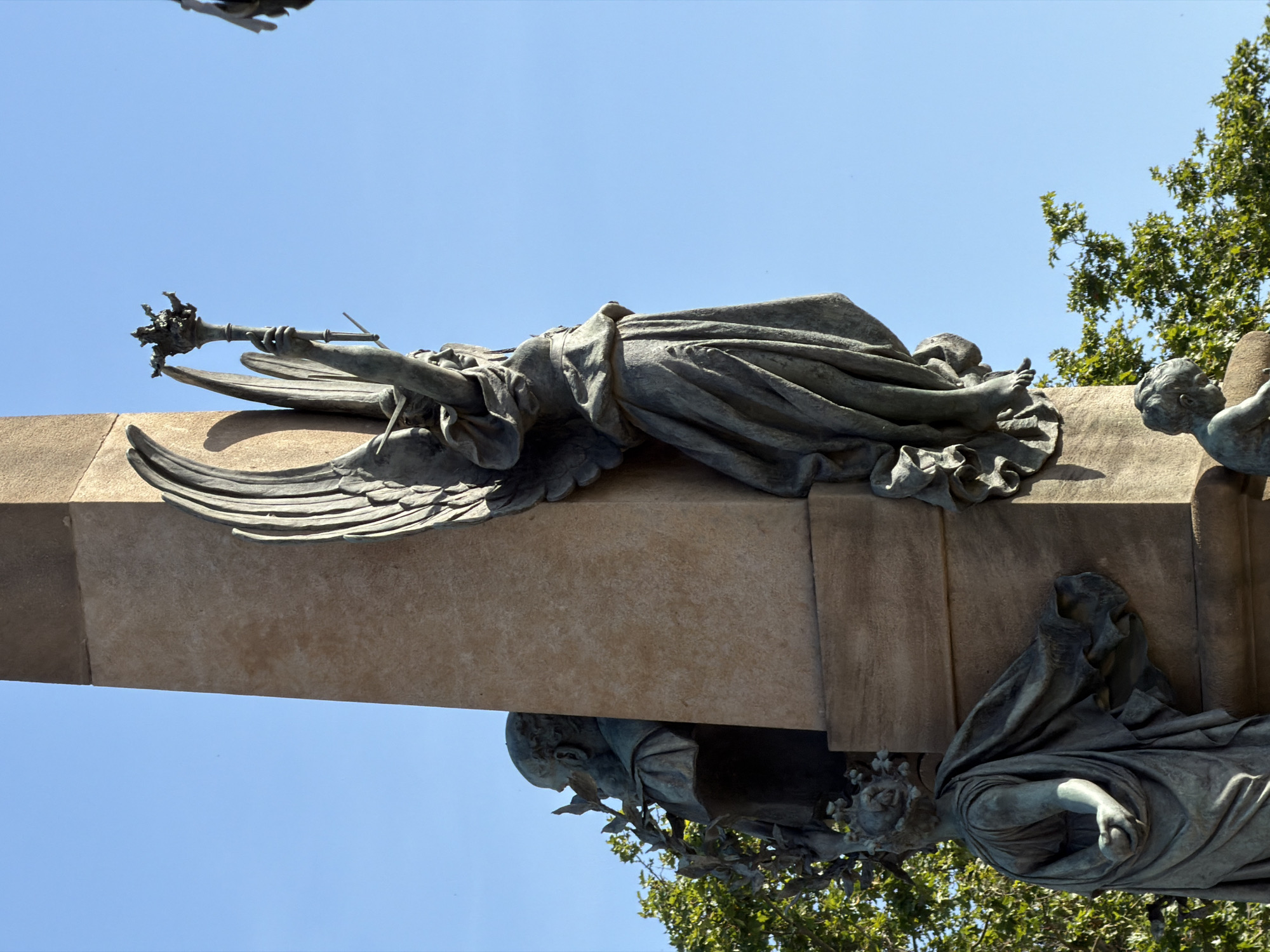 This is a medium shot of the Monument to Rafael Casanova in Barcelona, Spain. The monument, made of stone and bronze, is an obelisk with several allegorical statues. In the foreground, a bronze statue of an angel with outstretched wings holds a scepter topped with a crown. To the left, a bust of a man with a laurel wreath is partially visible. Below the angel, a young child's bronze statue is seated. The background is a clear blue sky with some green foliage visible to the left and lower right.